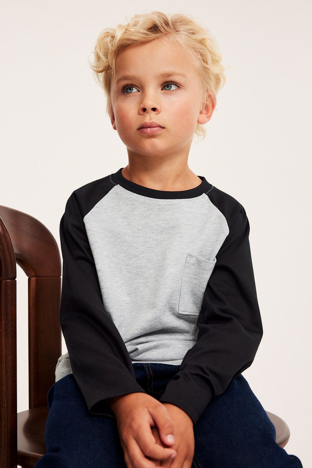 Framifrån: ett barn i en grå och svart långärmad raglantröja med bröstficka och mörkblå jeans.