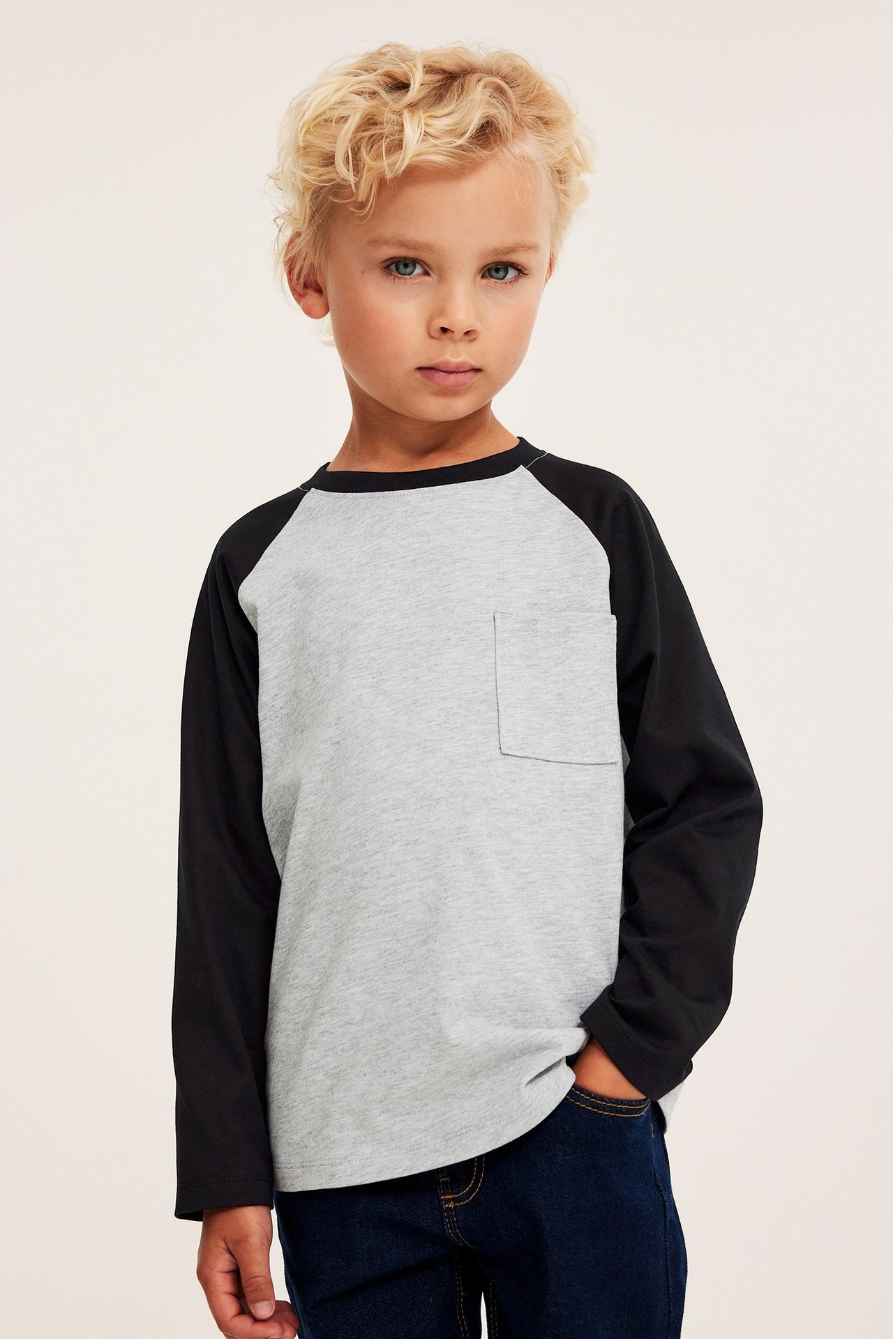 Widok z przodu: Dziecko w szaro-czarnej bawełnianej koszulce raglanowej z długim rękawem i kieszonką. Ciemnoniebieskie dżinsy.