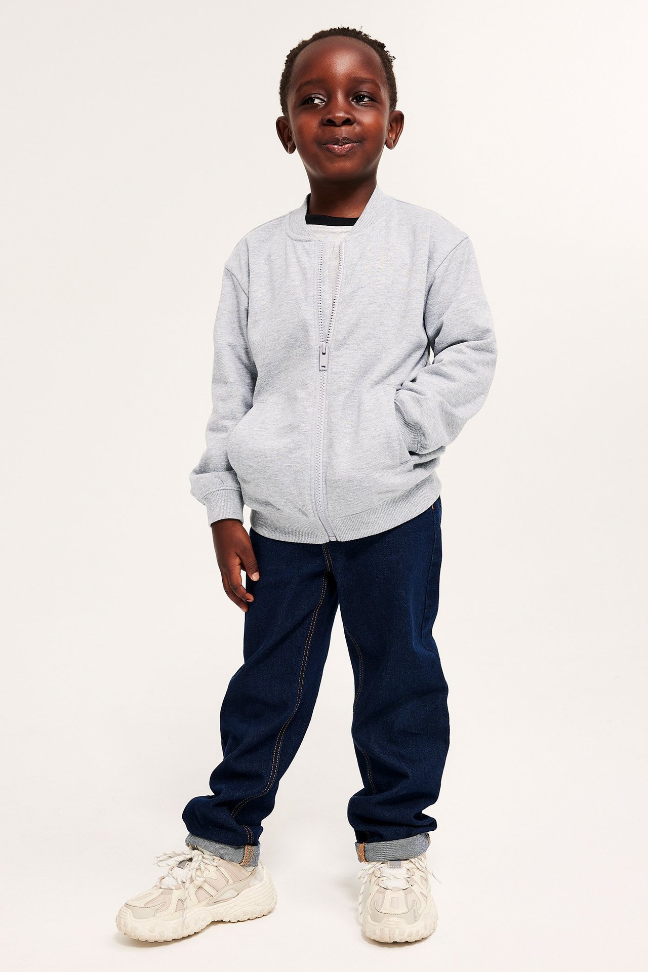 Edestä: Lapsi vaaleanharmaa vetoketjullinen neuletakki, tummansiniset farkut ja vaaleat lenkkarit.