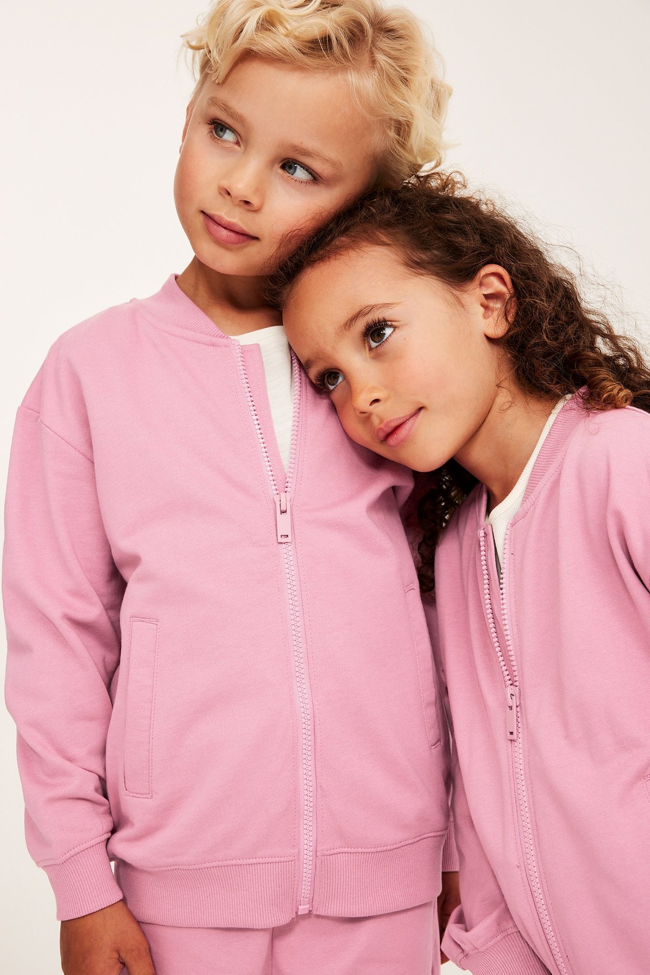 Lähikuva edestä kahdesta lapsesta yllään vaaleanpunaiset puuvillaiset vetoketjulliset neuletakit taskuilla ja valkoisilla paidoilla.