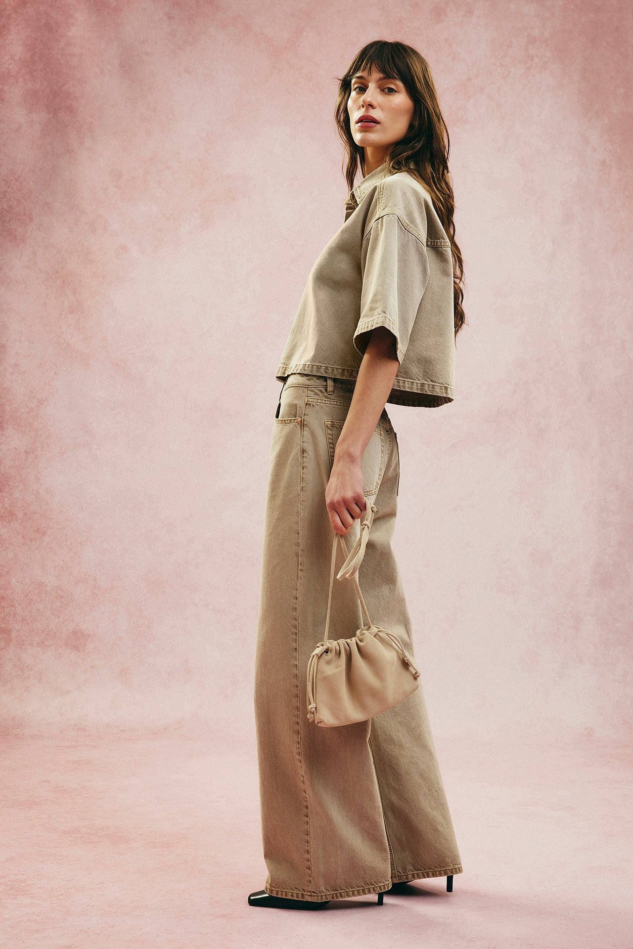 Sidovy: Kvinna i ljusbruna vida jeans med hög midja, matchande kort topp, håller en liten beige väska.