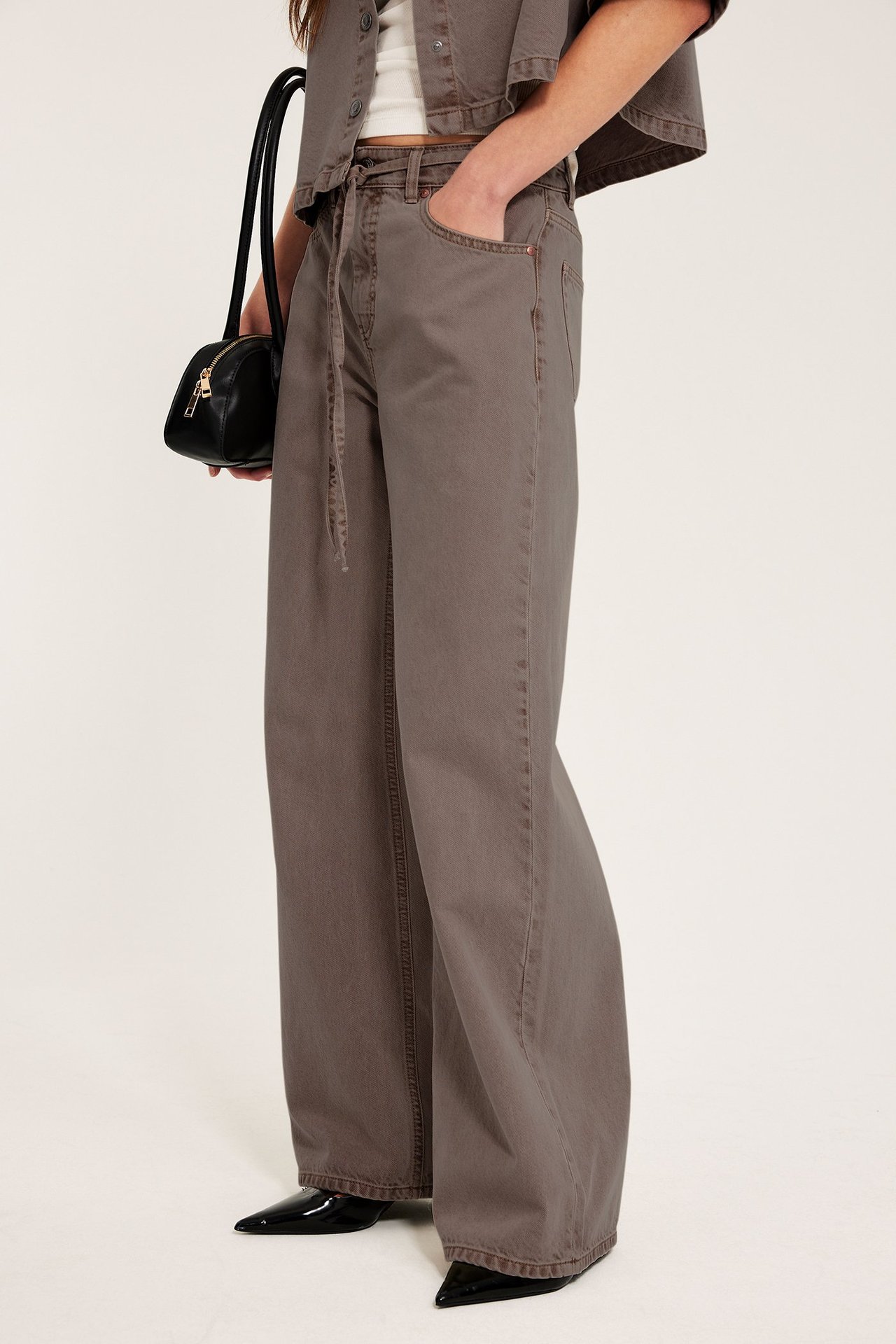 Kvinne i lysebrune wide-leg jeans med knytebelte, kort brun skjorte, svart veske og hæler.