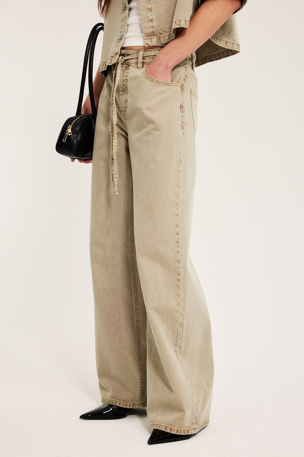 Nainen vaaleanruskeat korkeavyötäröiset leveät farkut solmimisvyöllä, lyhyt takki, pitää mustaa käsilaukkua.