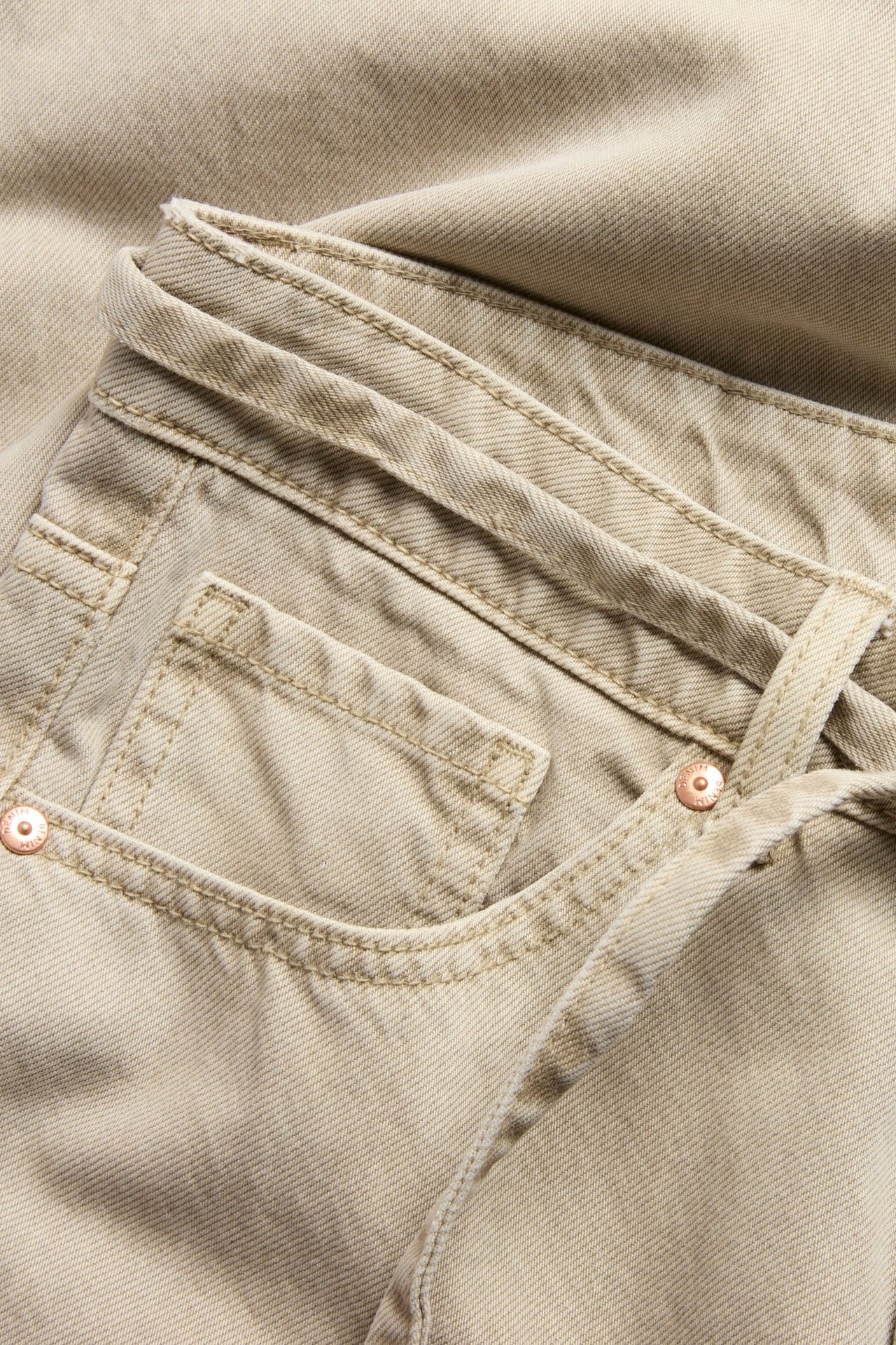 Lähikuva vaaleanruskeista korkeavyötäröisistä leveälahkeisista twill-farkuista etutaskulla ja vyöllä.