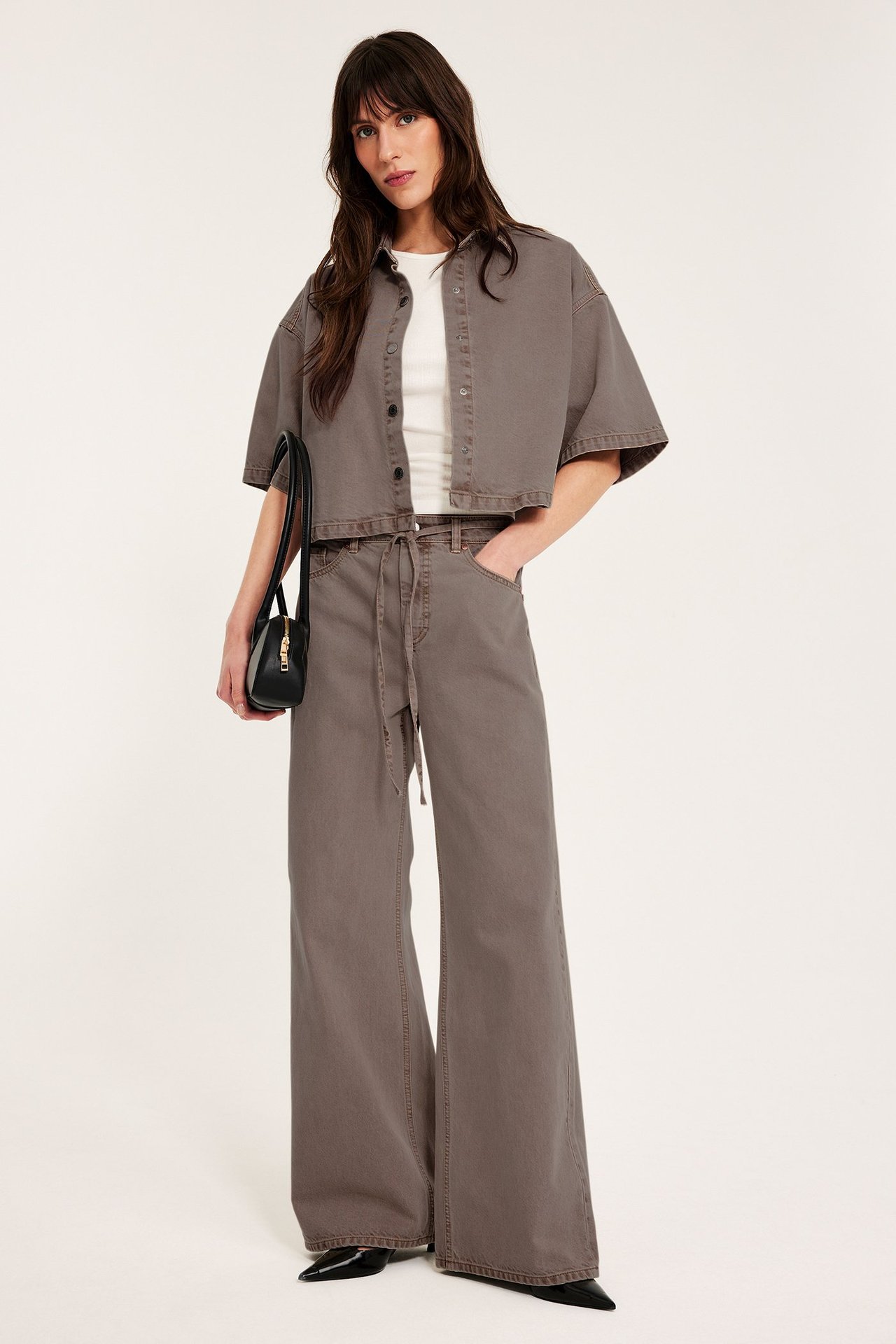 Kobieta od przodu w jasnobrązowych szerokich jeansach z wysokim stanem, pasującej krótkiej koszuli i czarnej torebce.