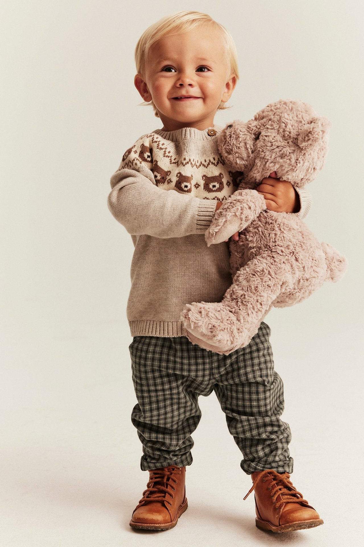 Edestä: vauva ruskeissa ruudullisissa kudotuissa housuissa, beigessä nallepuserossa ja ruskeissa kengissä, pitäen nallea.