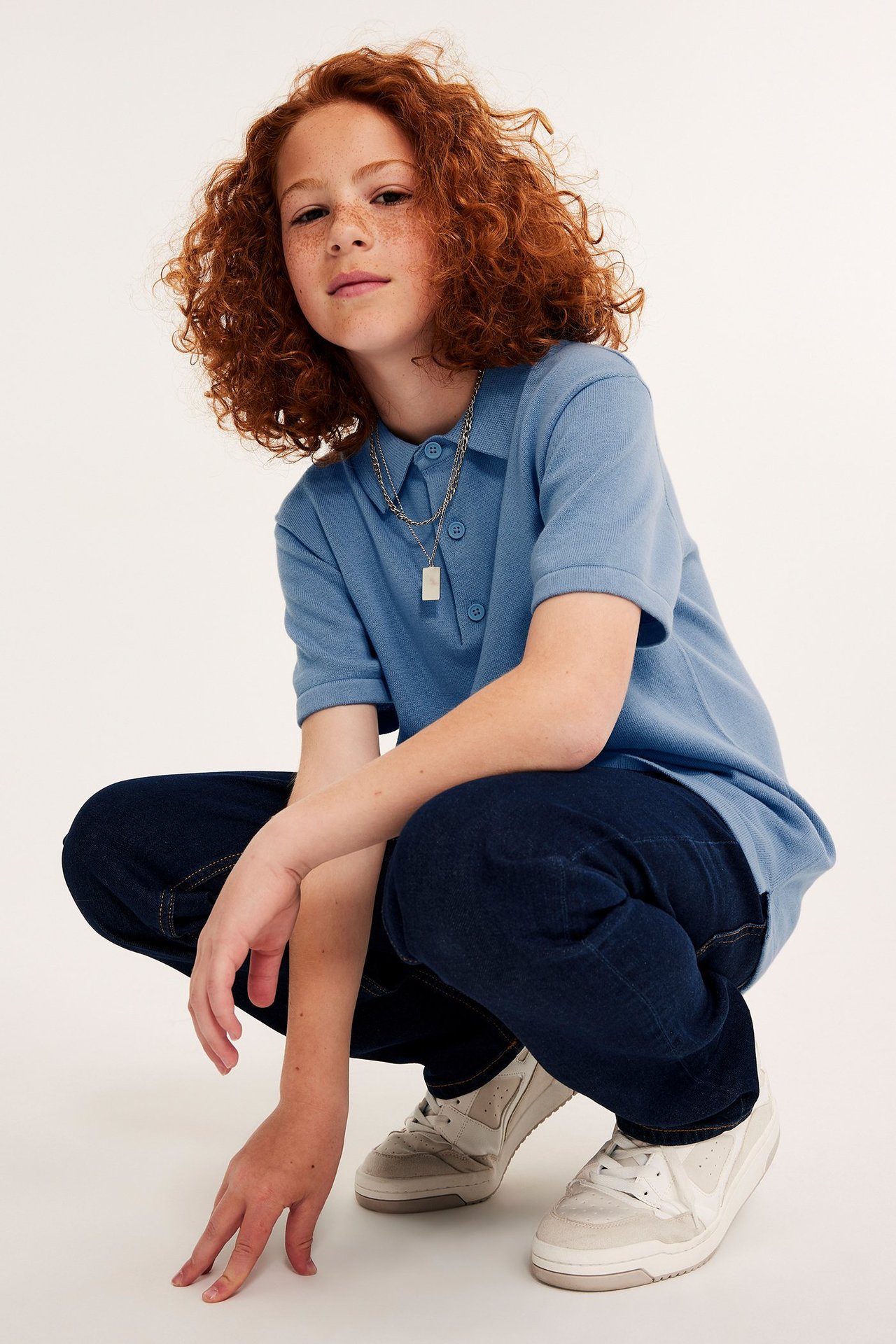 Widok z przodu: Dziecko w niebieskiej, drobno dzianej koszulce polo, ciemnoniebieskich dżinsach, białych trampkach i naszyjniku.