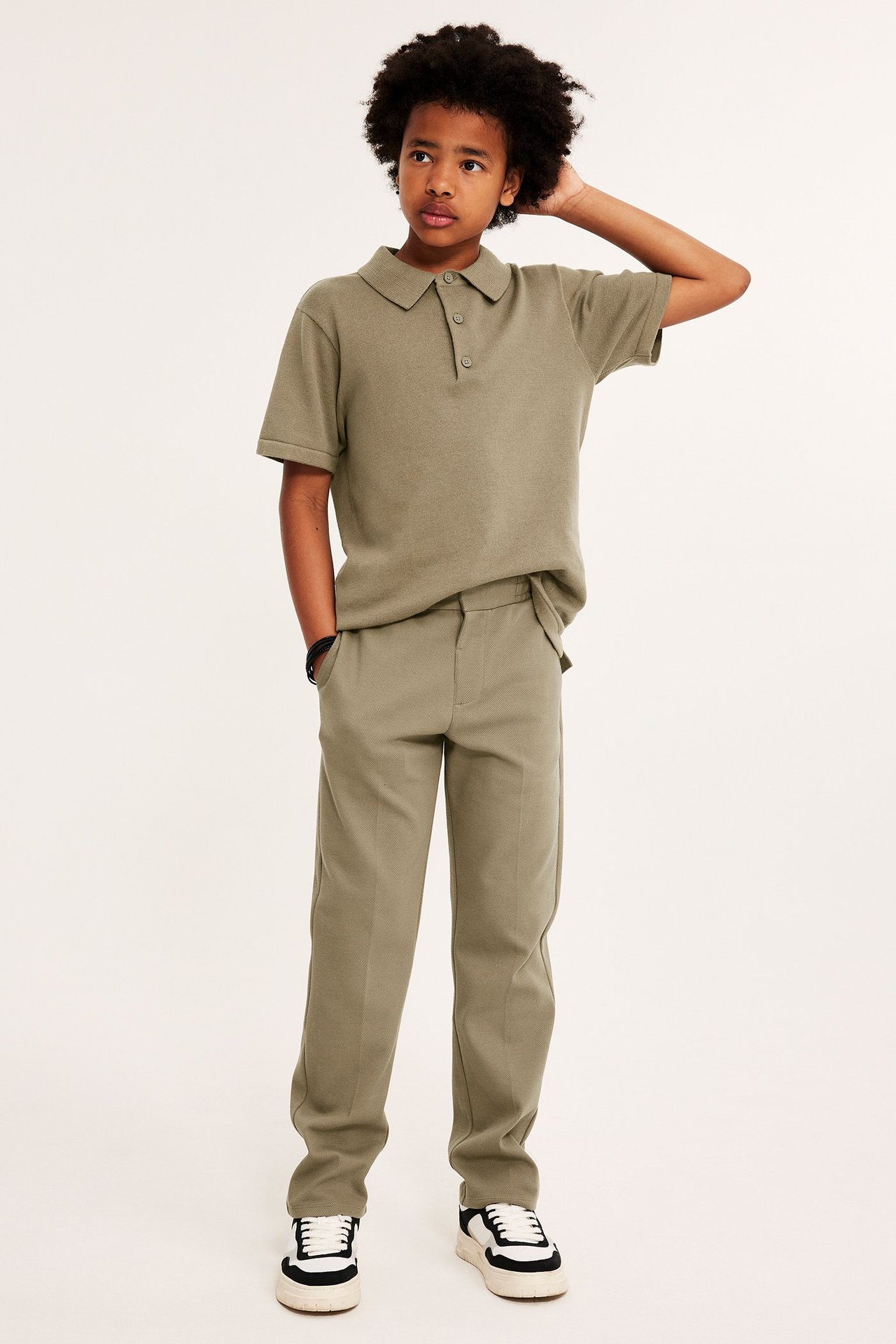 Widok z przodu dziecka w ciemnobeżowej, drobno dzianej koszulce polo, pasujących spodniach i czarno-białych sneakersach.