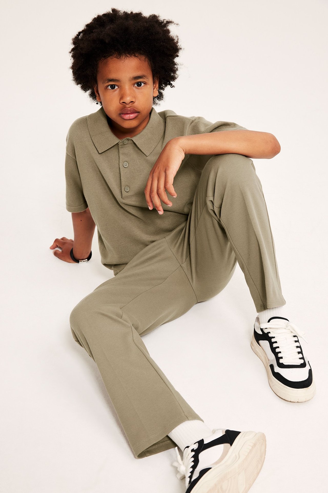 Widok z przodu: dziecko w ciemnobeżowej, dzianej koszulce polo, pasujących spodniach i czarno-białych sneakersach.