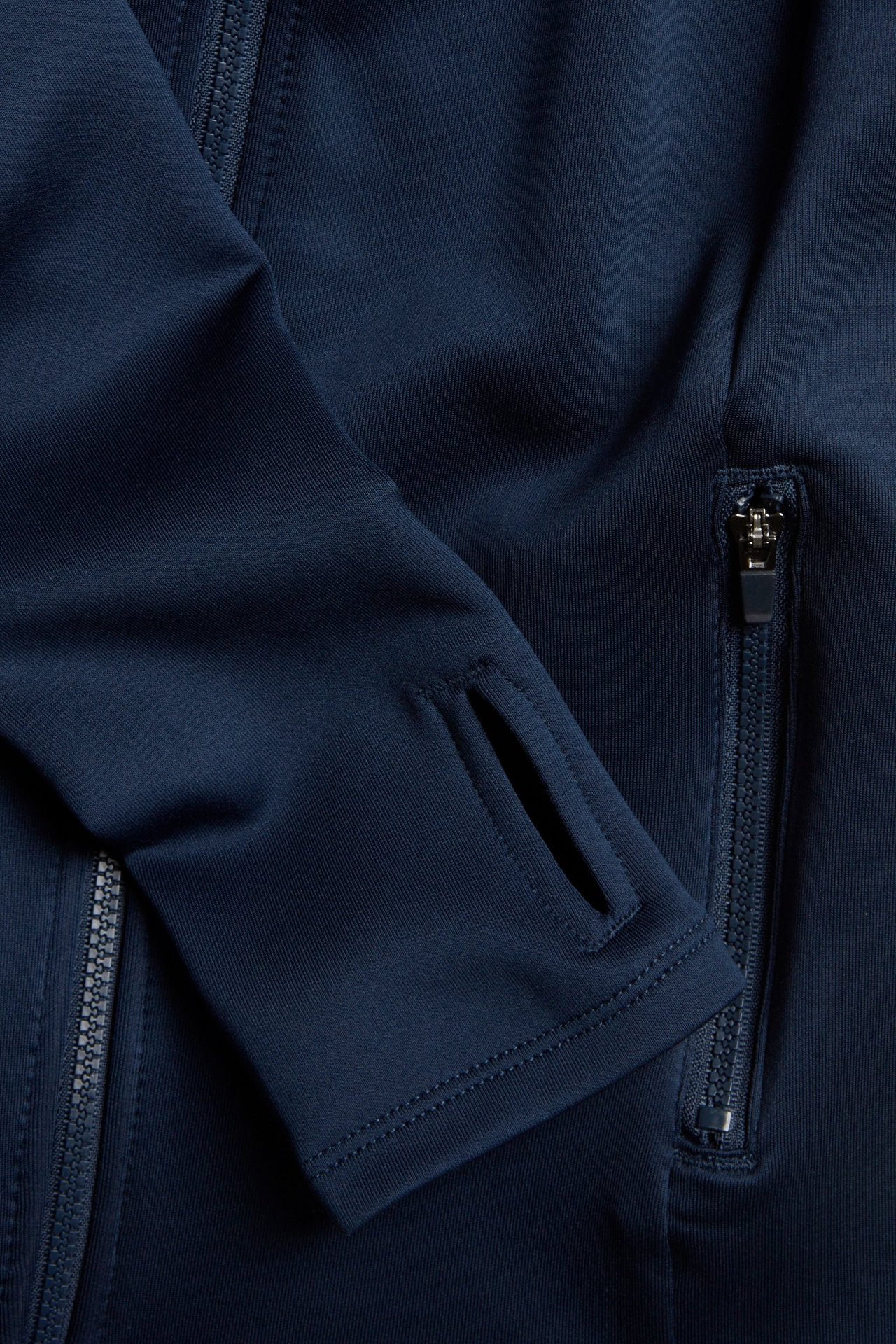 Lähikuva tummansinisestä treenitakista, jossa on peukaloaukko hihassa ja vetoketjullinen tasku.