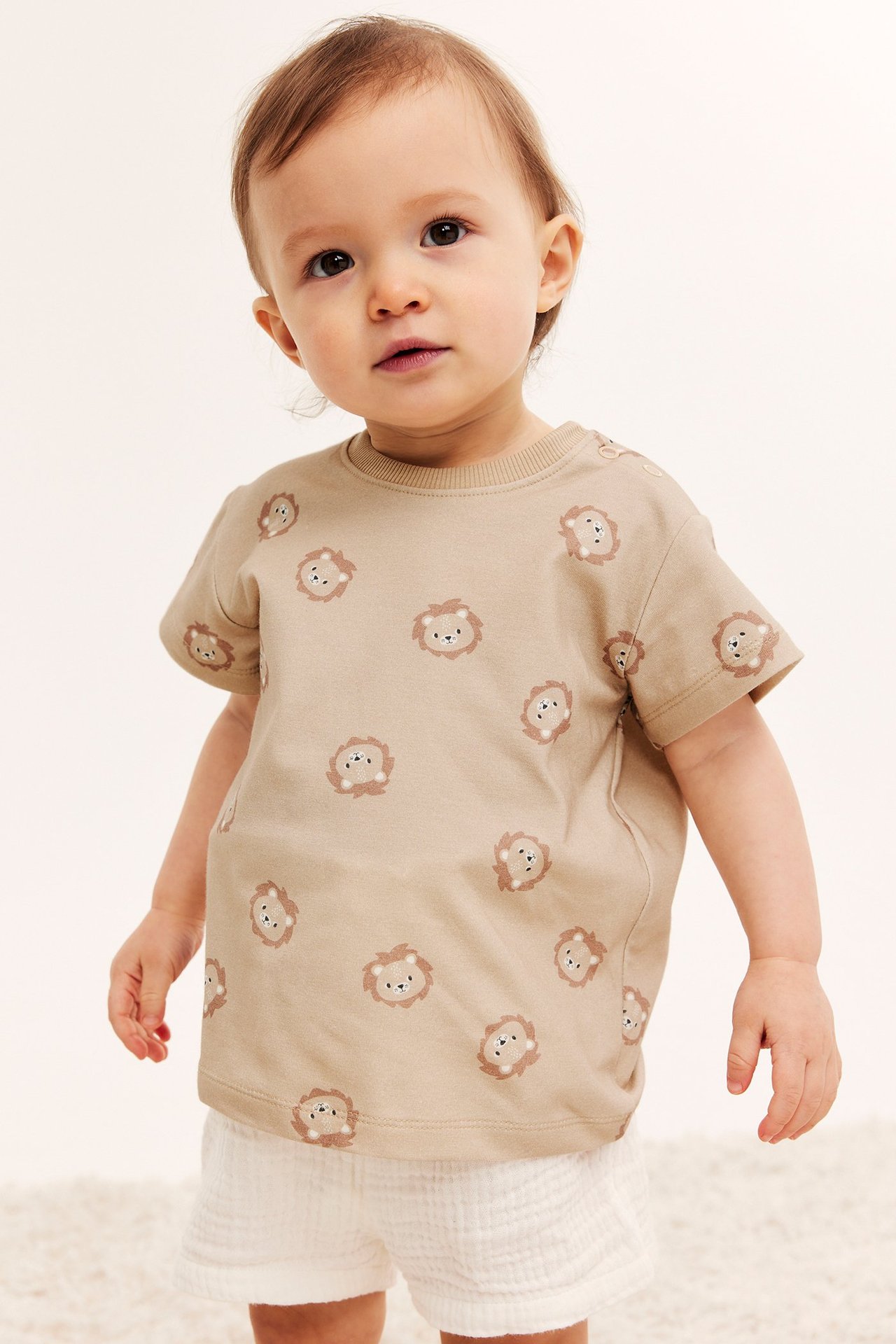 Forfra: baby i beige kortermet t-skjorte med løvetrykk, trykknapper og hvite krøllete shorts.