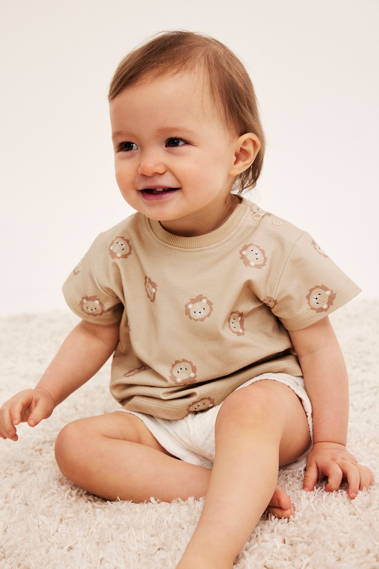 Widok z przodu uśmiechniętego dziecka w beżowej koszulce z lwem i białych szortach.