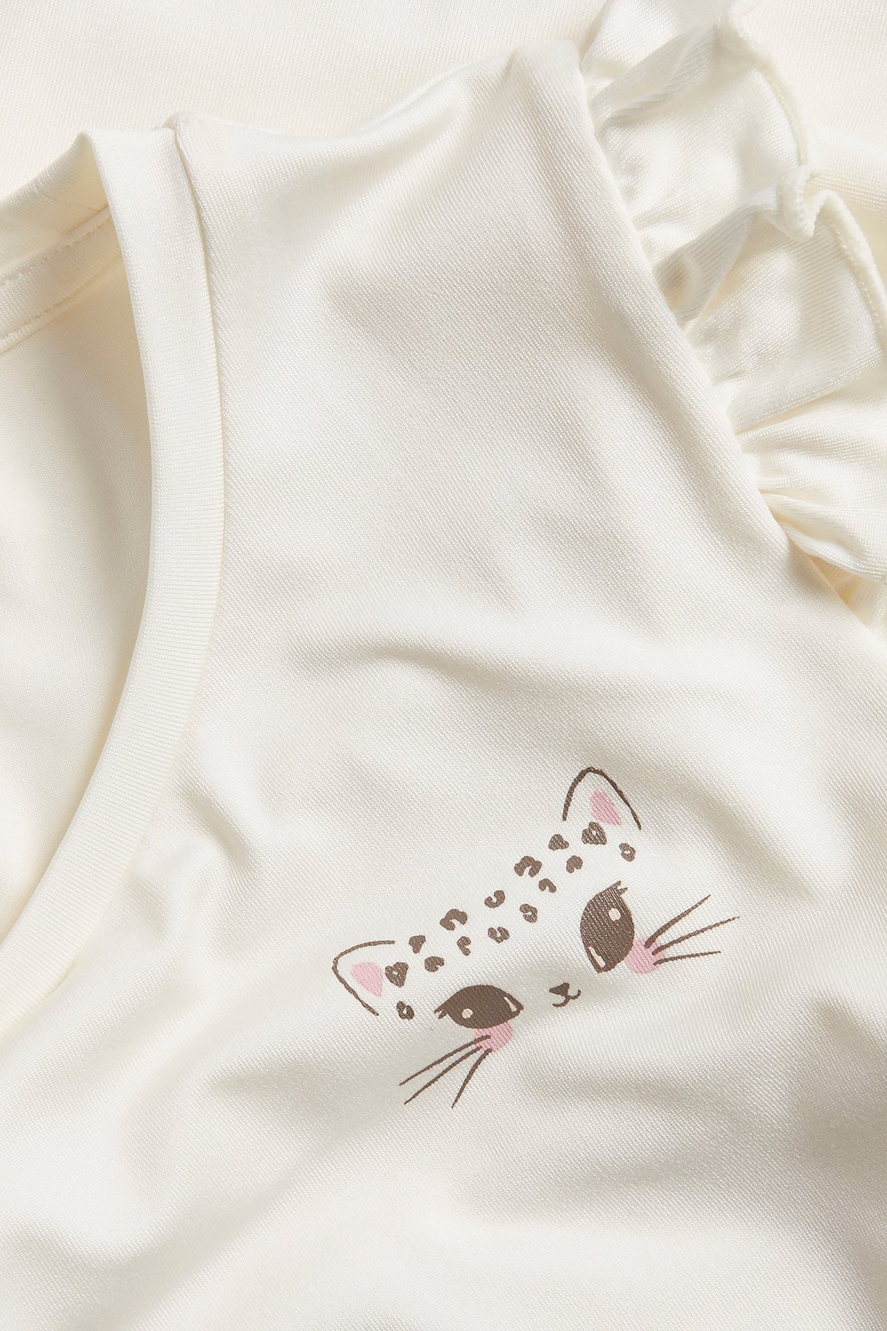 Närbild av en offwhite träningstopp för barn med volang på axeln och kattansikte-tryck.