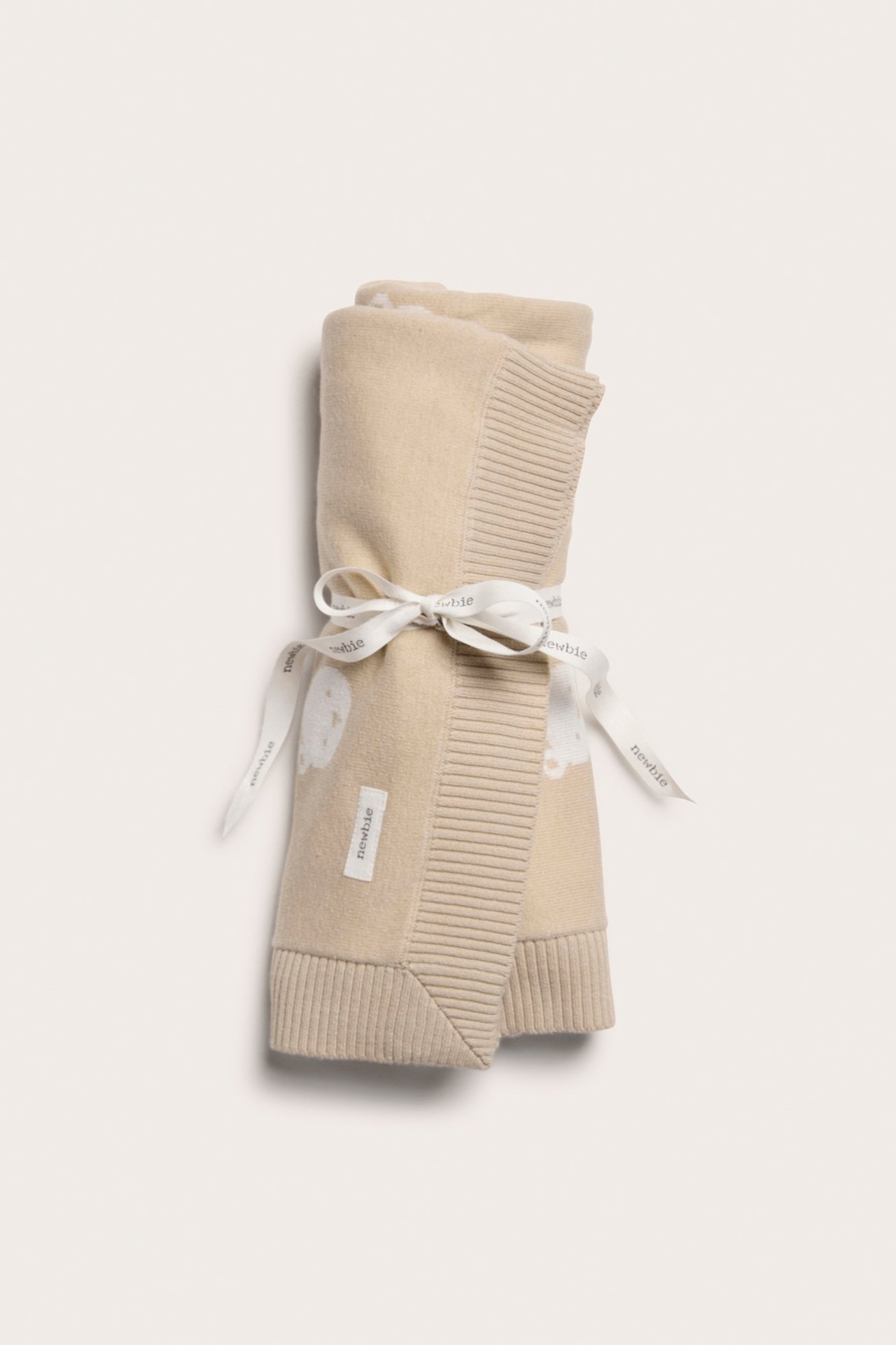 Framifrån, en rullad beige jacquardstickad bomullsfilt med nallemönster, knuten med ett vitt band.