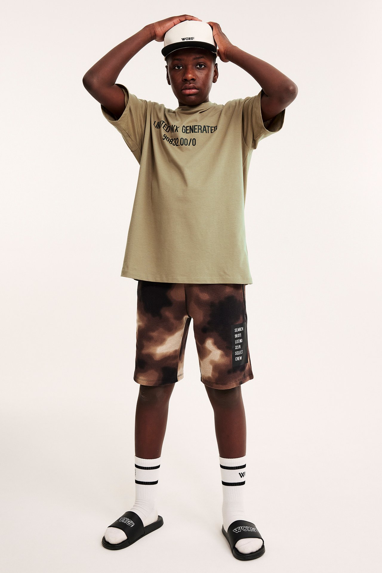 Dziecko w oversize'owym khaki t-shircie z nadrukiem, brązowych szortach tie-dye, białych skarpetkach, czarnych klapkach, trzyma białą czapkę.