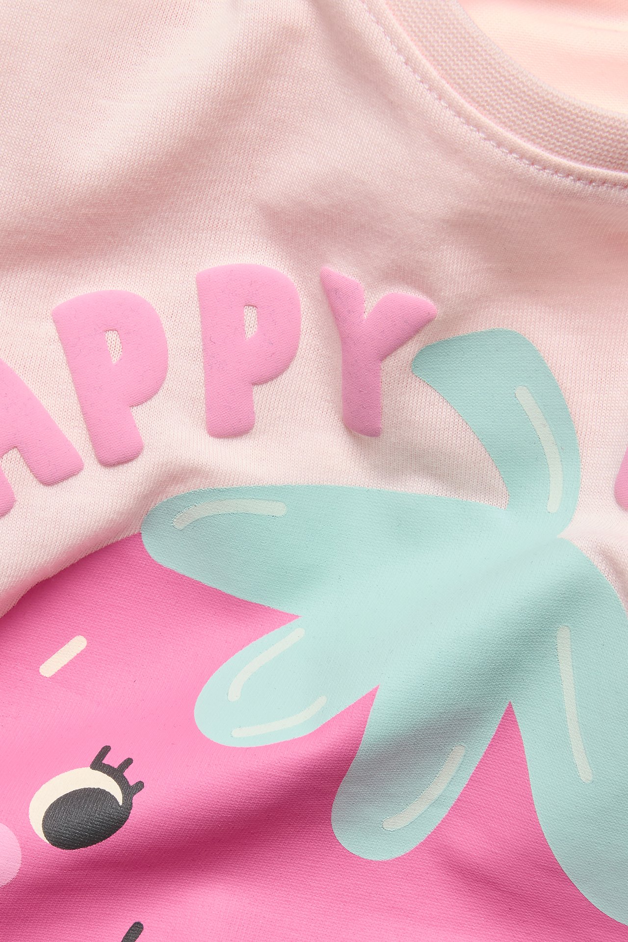 Närbild av en rosa bomulls-t-shirt för barn med upphöjt "HAPPY"-tryck och färgglad grafik.