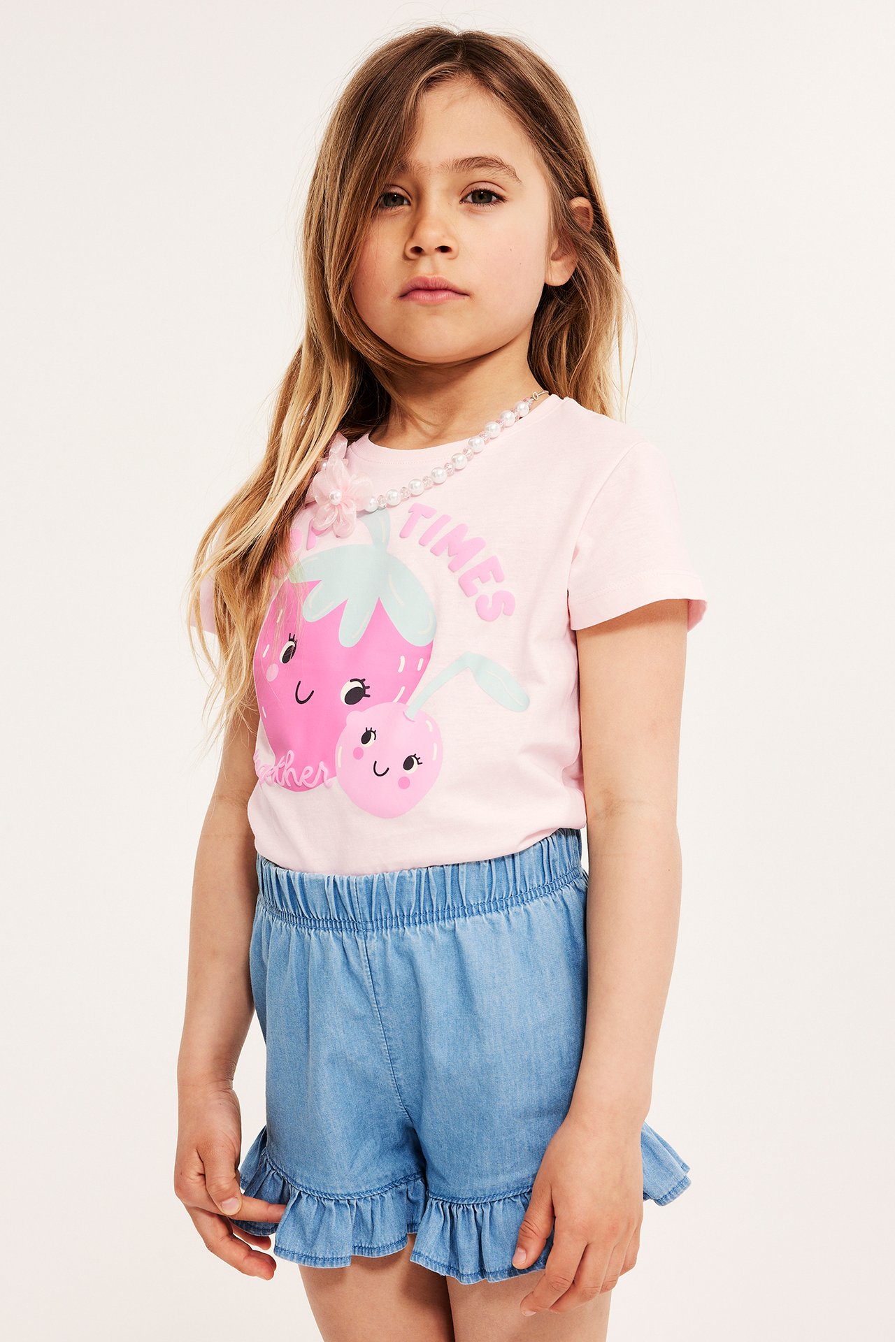 Framifrån: Barn i rosa bomulls-t-shirt med frukttryck, ljusblå volangshorts och pärlhalsband.