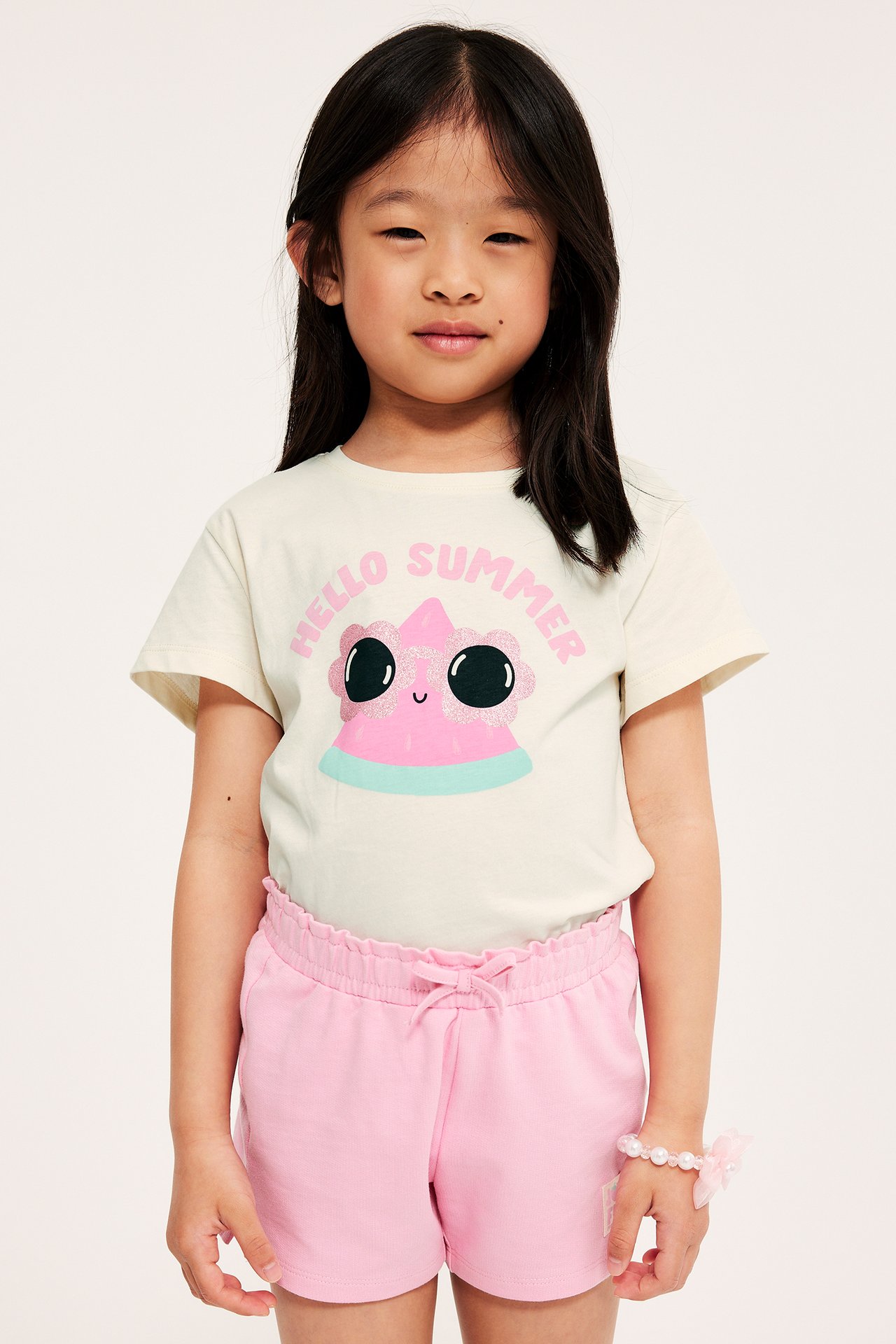 Framifrån: Barn i offwhite bomulls-t-shirt med rosa glitter-glass-tryck, rosa shorts och pärlarmband.
