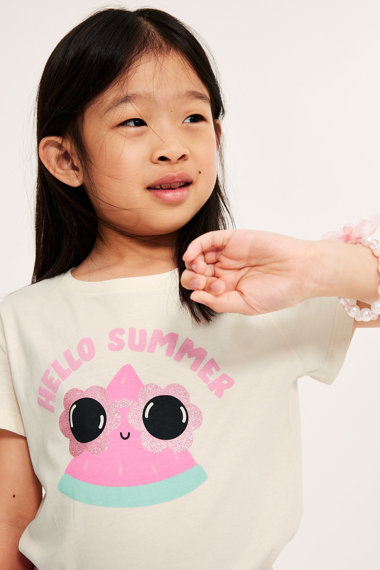 Närbild på ett barn i en offwhite bomulls-T-shirt med rosa vattenmelon-tryck och ett pärlarmband.