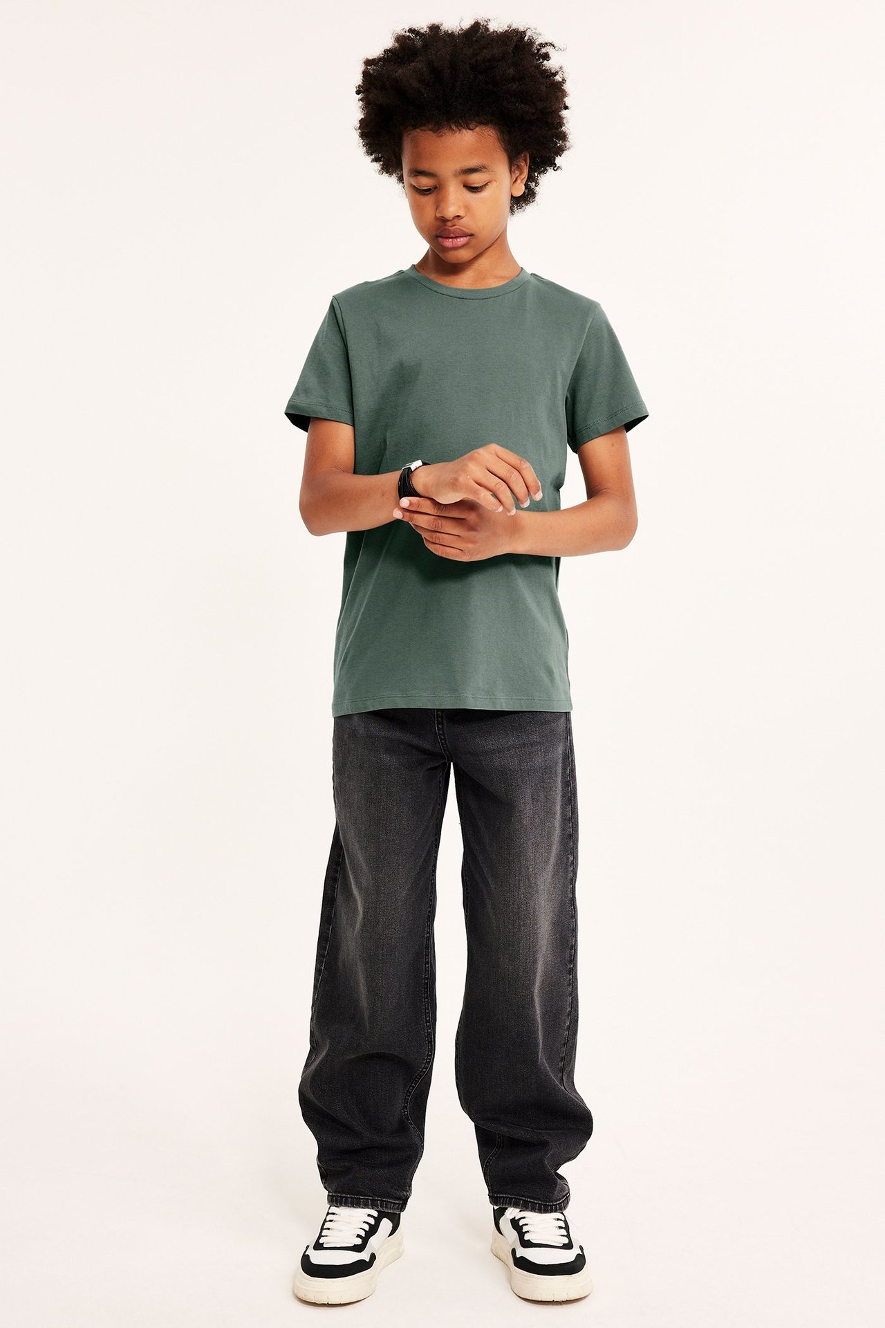 Hel frontvy av ett barn i en enfärgad khakigrön bomulls-t-shirt, mörkgrå vida jeans och sneakers.