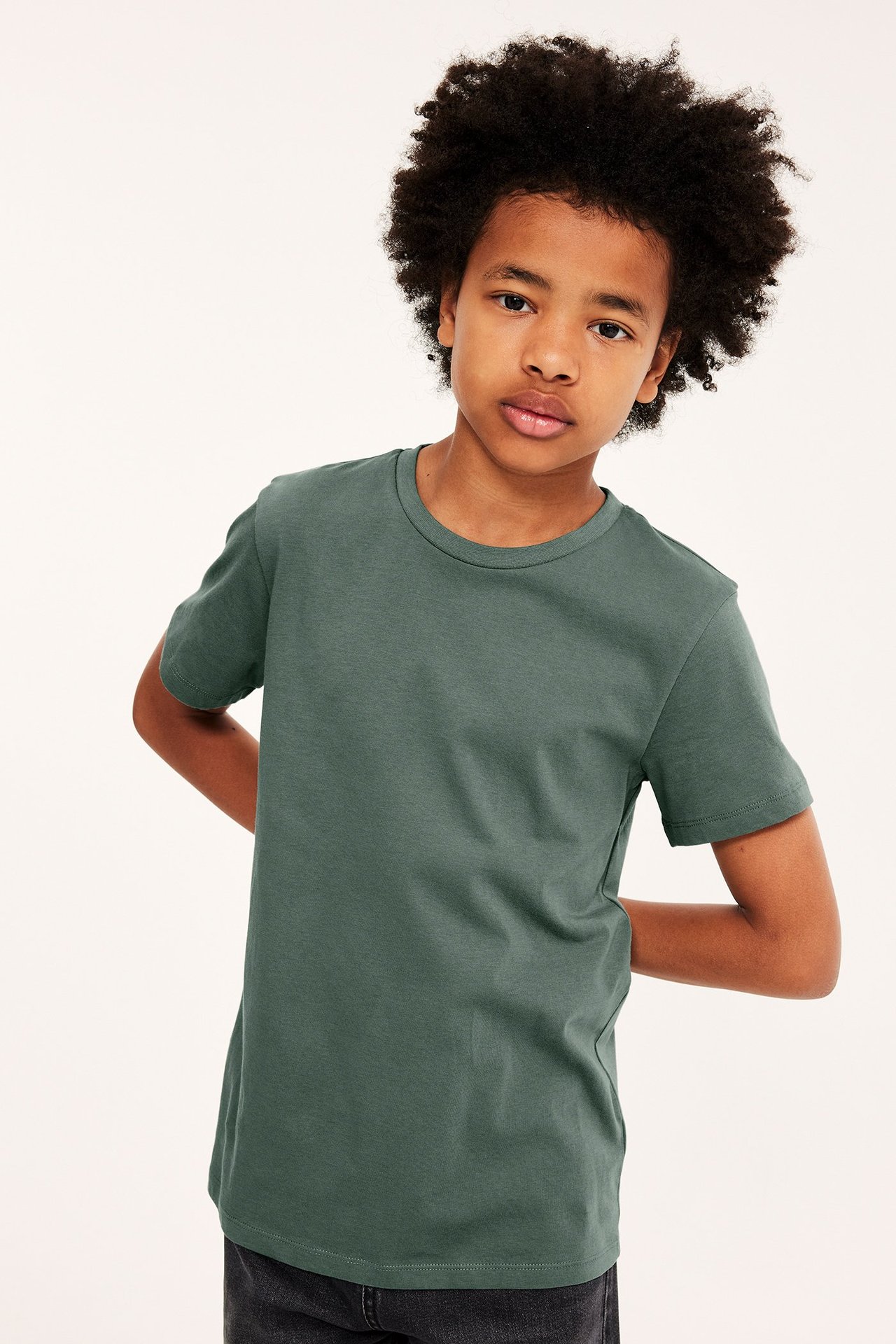 Frontvy av ett barn i en enfärgad khakigrön kortärmad t-shirt i bomull och mörka jeans.
