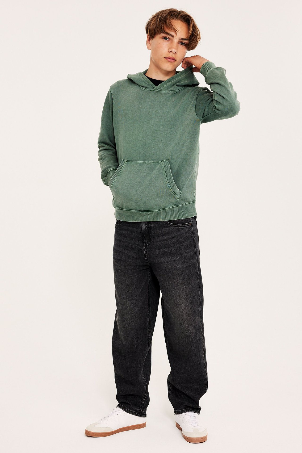 Framifrån: Barn i tvättad khakigrön bomullshoodie med kängruficka, mörkgrå jeans och vita sneakers.