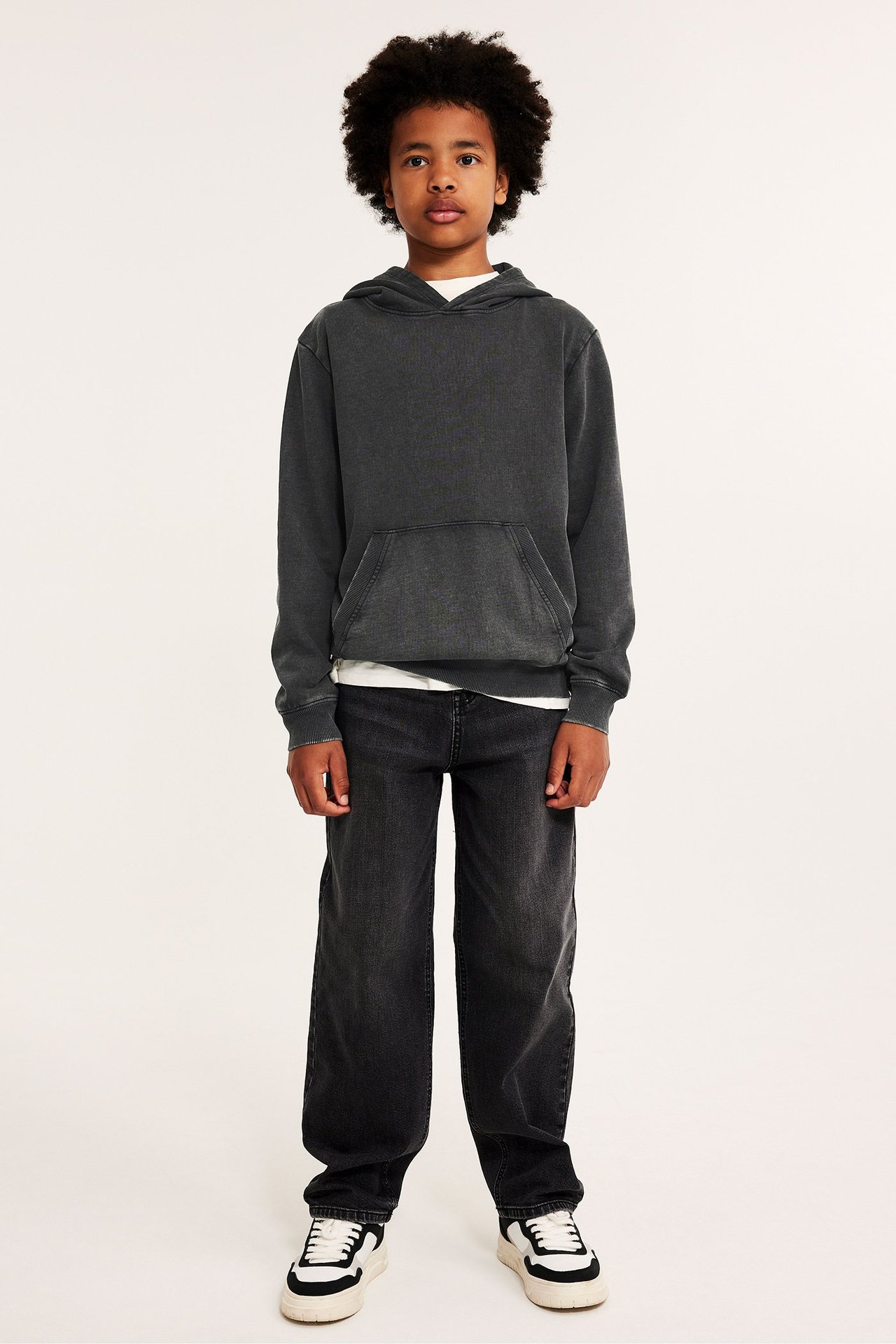 Framifrån: Kid i mörkgrå bomullshoodie, vit t-shirt, svarta jeans och svartvita sneakers.