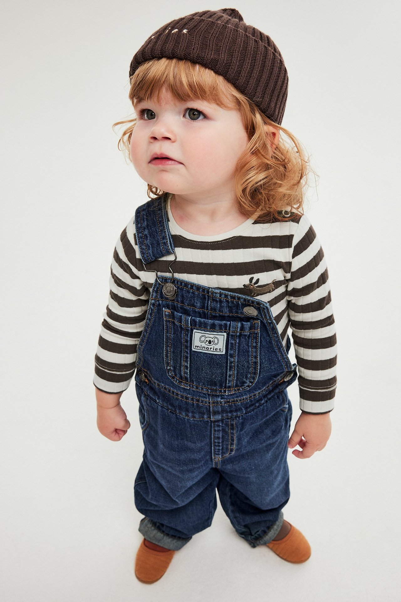 Edestä: vauva yllään ruskeavalkoraidallinen ribbibody, siniset farkkuhaalarit, ruskea pipo ja kengät.