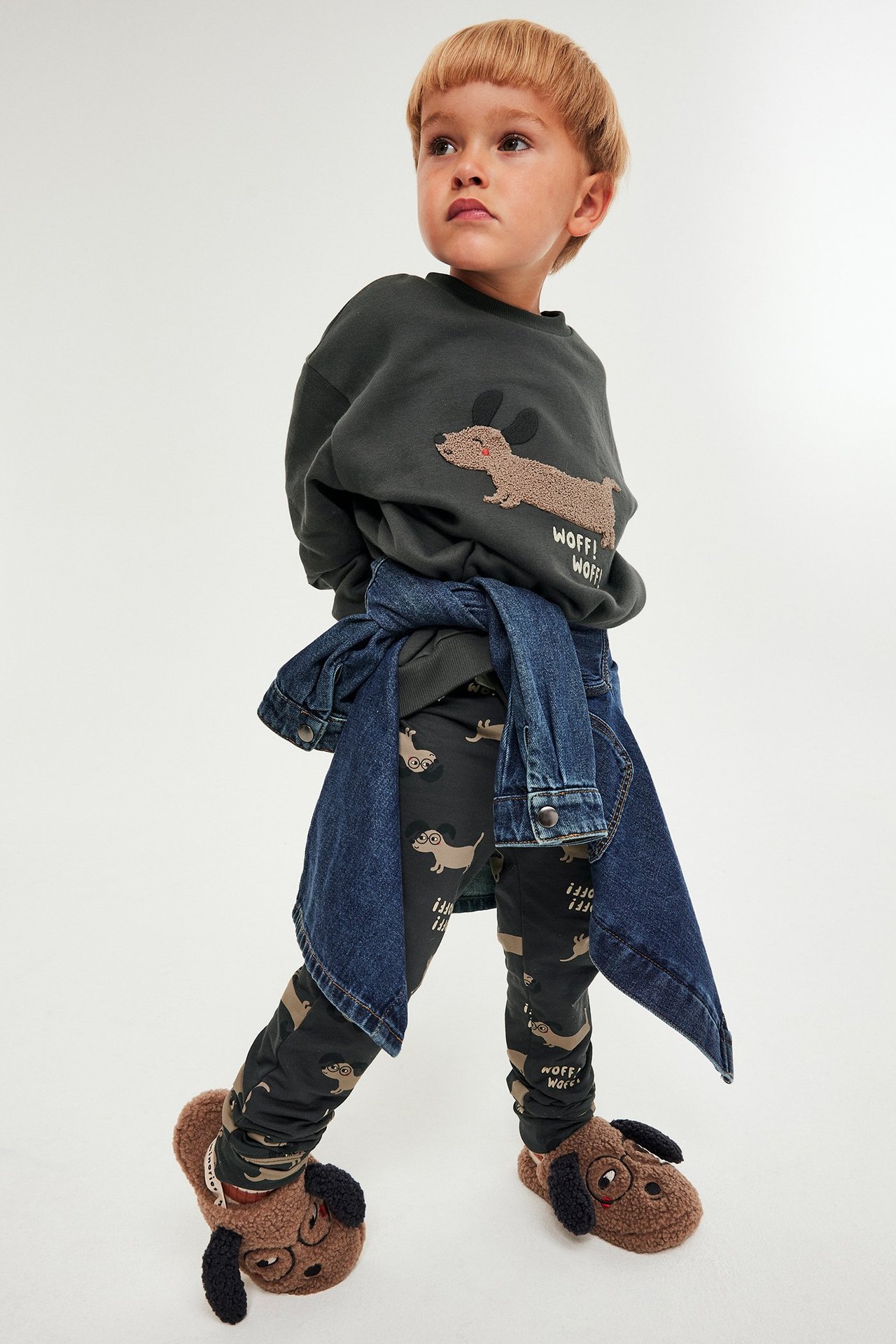 Lapsi mustassa koira-aplikoidussa collegepaidassa, tummissa leggingseissä, farkkutakissa ja koiratossuissa, edestä päin.
