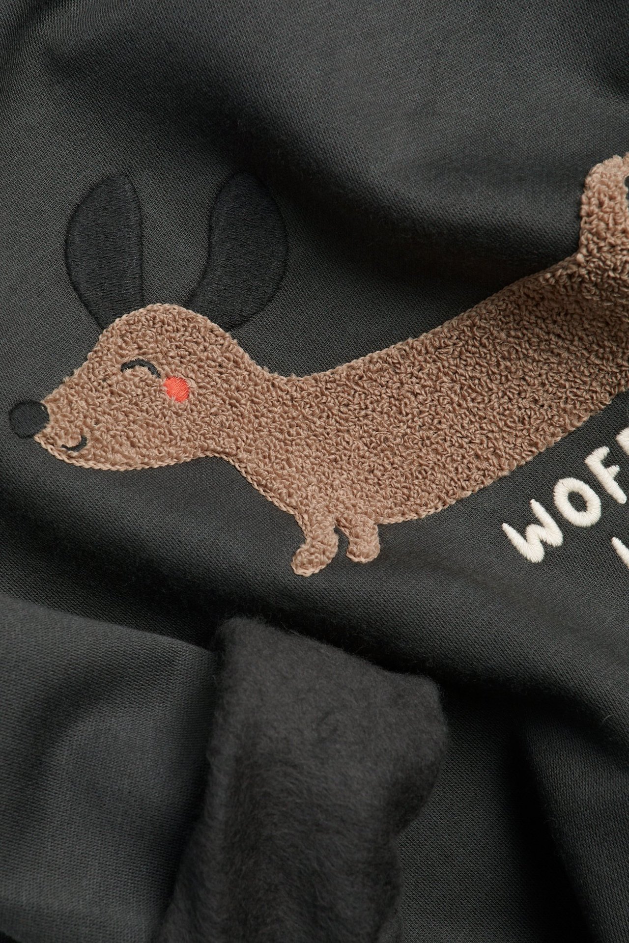 Närbild på en svart sweatshirt med brun frottéapplikation av tax och vit "WOF"-brodyr. Borstad insida synlig.