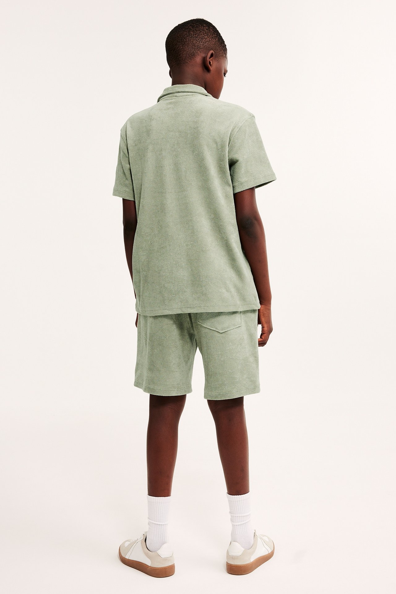 Bakifrån: ett barn i mörkgröna frottéjersey shorts med bakficka, matchande tröja, vita strumpor och sneakers.