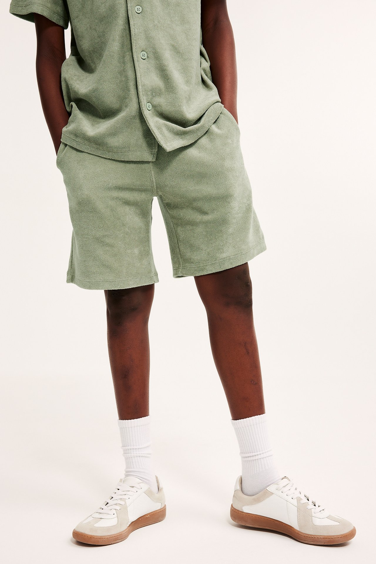 Framifrån, barn i mörkgröna frottéshorts, matchande skjorta, vita strumpor och vita/beige sneakers.
