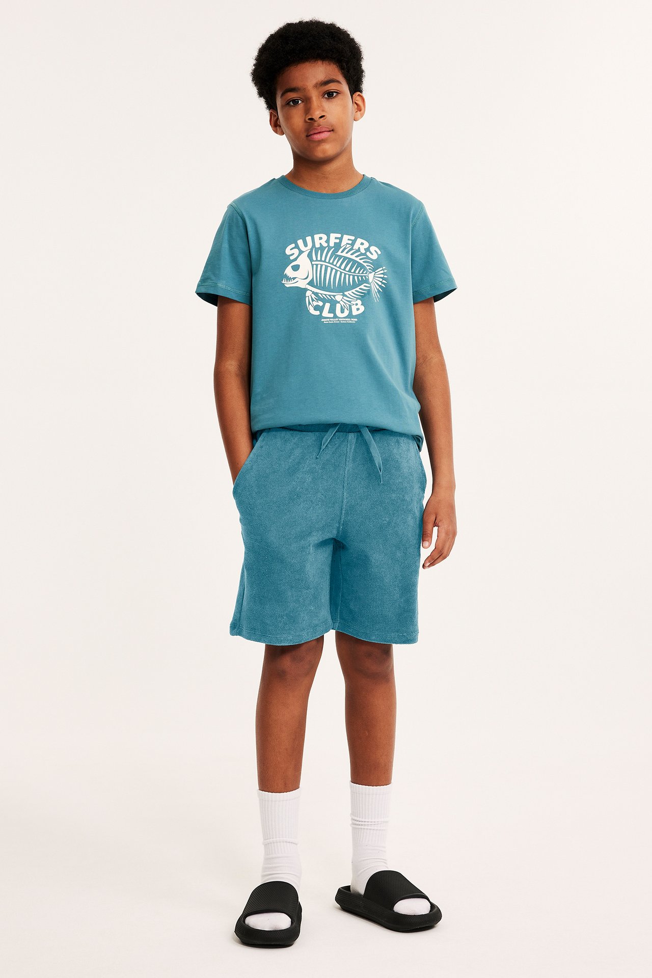 Barn i grønne frottéshorts med snøring, turkis t-skjorte med trykk, hvite sokker og svarte sandaler, frontbilde.