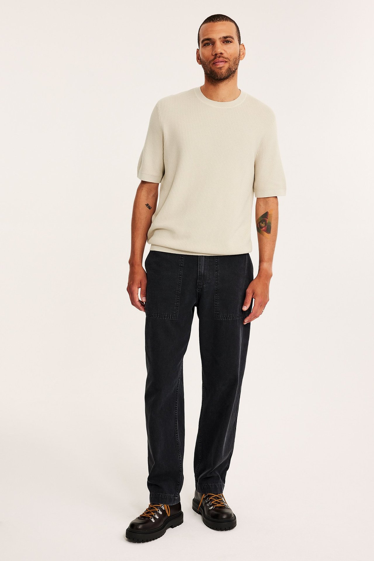 Edestä: mies yllään vaaleanbeige vohvelineulottu lyhythihainen puuvillaneule, tummanharmaat housut ja ruskeat nauhakengät.