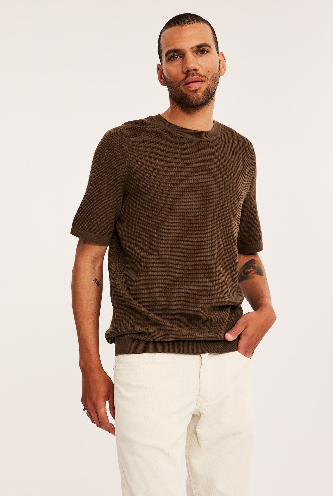 Edestä: mies ruskeassa vohvelineulotussa lyhythihaisessa neuleessa ja vaaleissa housuissa, käsi taskussa.