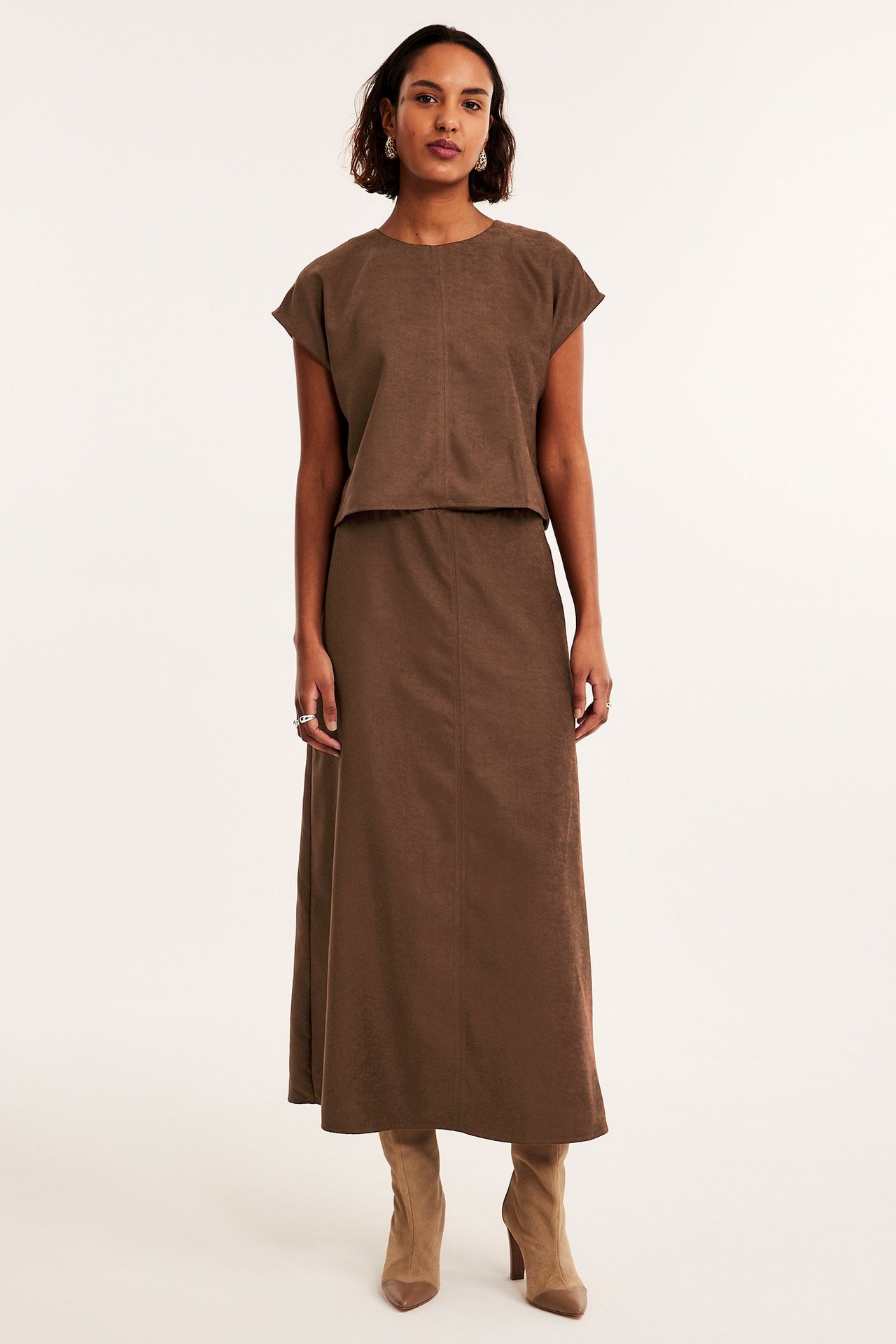 Frontbilde av kvinne i brun fuskemokka croptopp, matchende langt skjørt og lysebrune støvler.