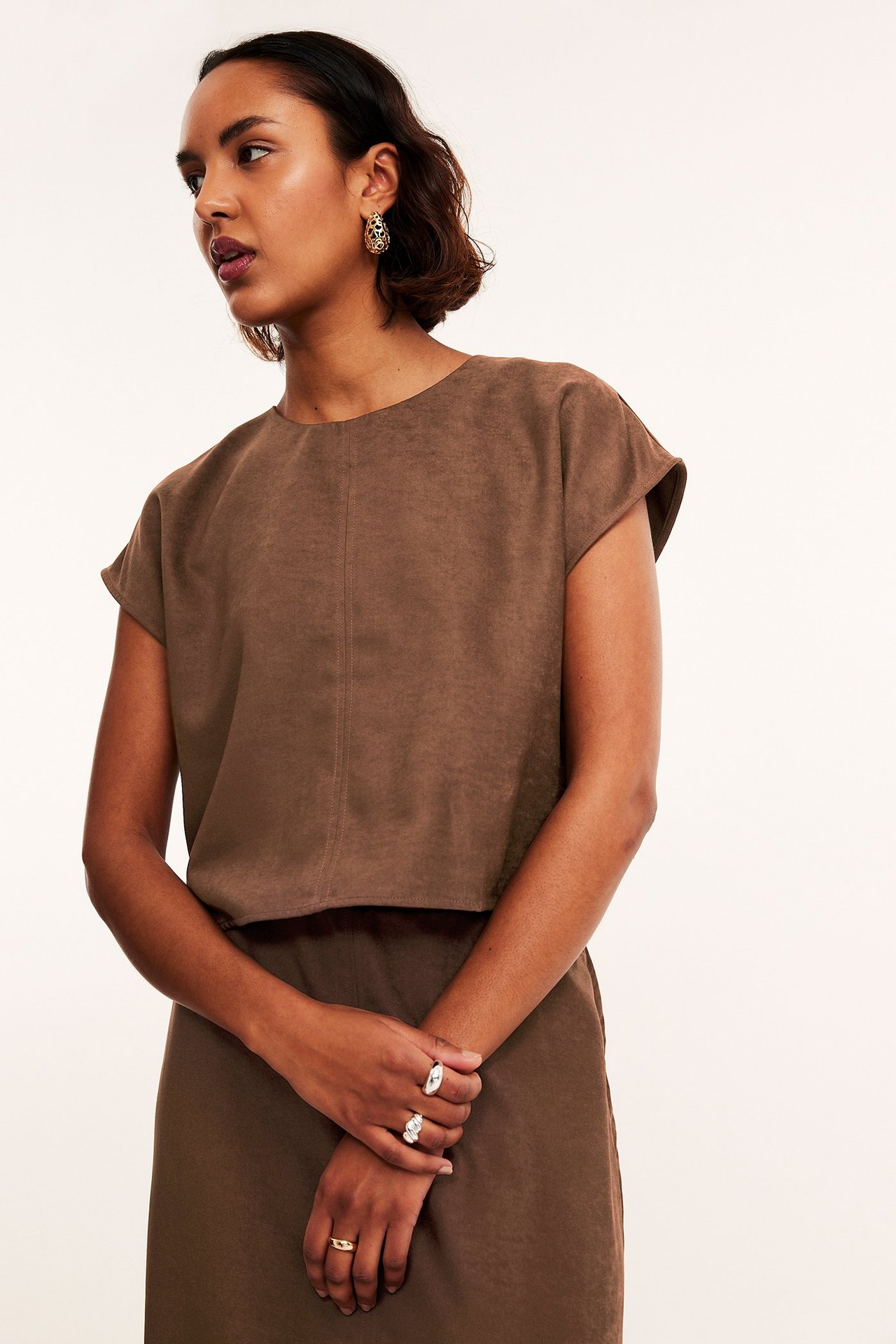 Widok z przodu/boku kobiety w brązowym krótkim topie z imitacji zamszu, brązowej spódnicy, złotych kolczykach i pierścionkach.