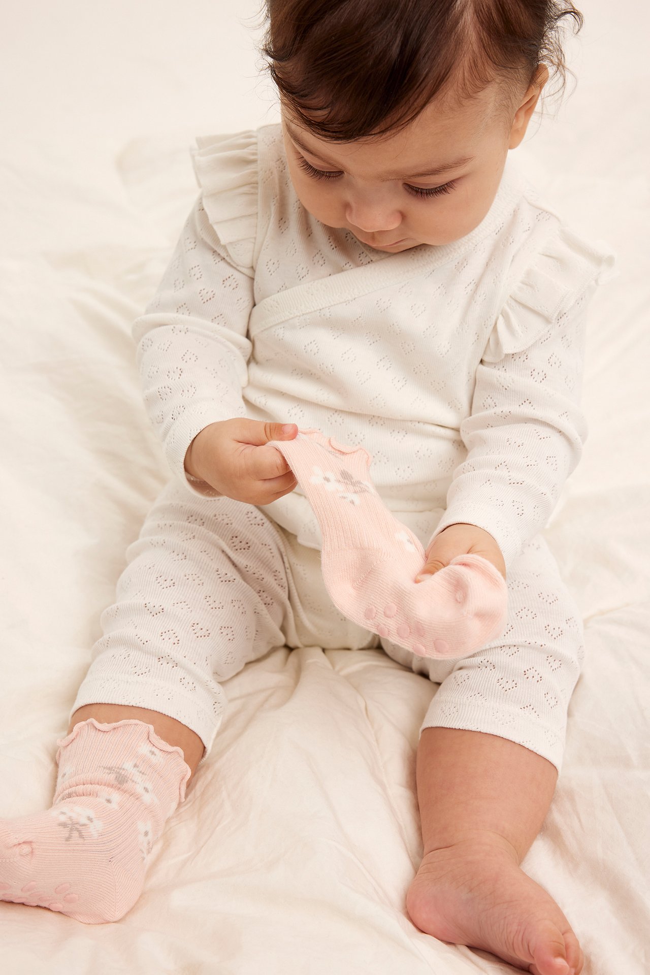 Framifrån: baby håller en ljusrosa blommig strumpa, bär en matchande strumpa och vit mönstrad outfit.