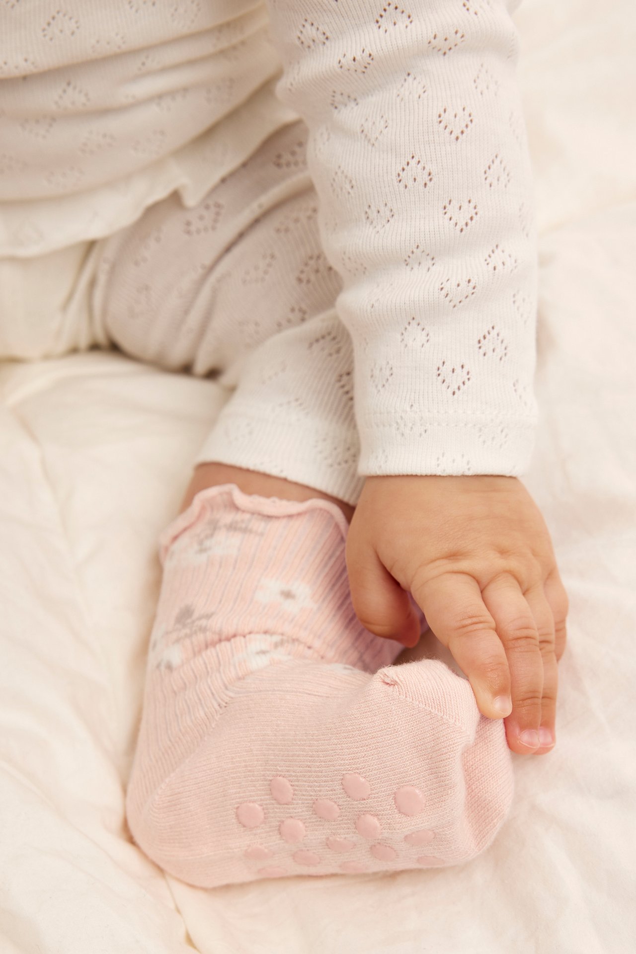 Närbild: Bebisens hand rör en rosa blommig strumpa med halkskydd, buren med vita mönstrade kläder.
