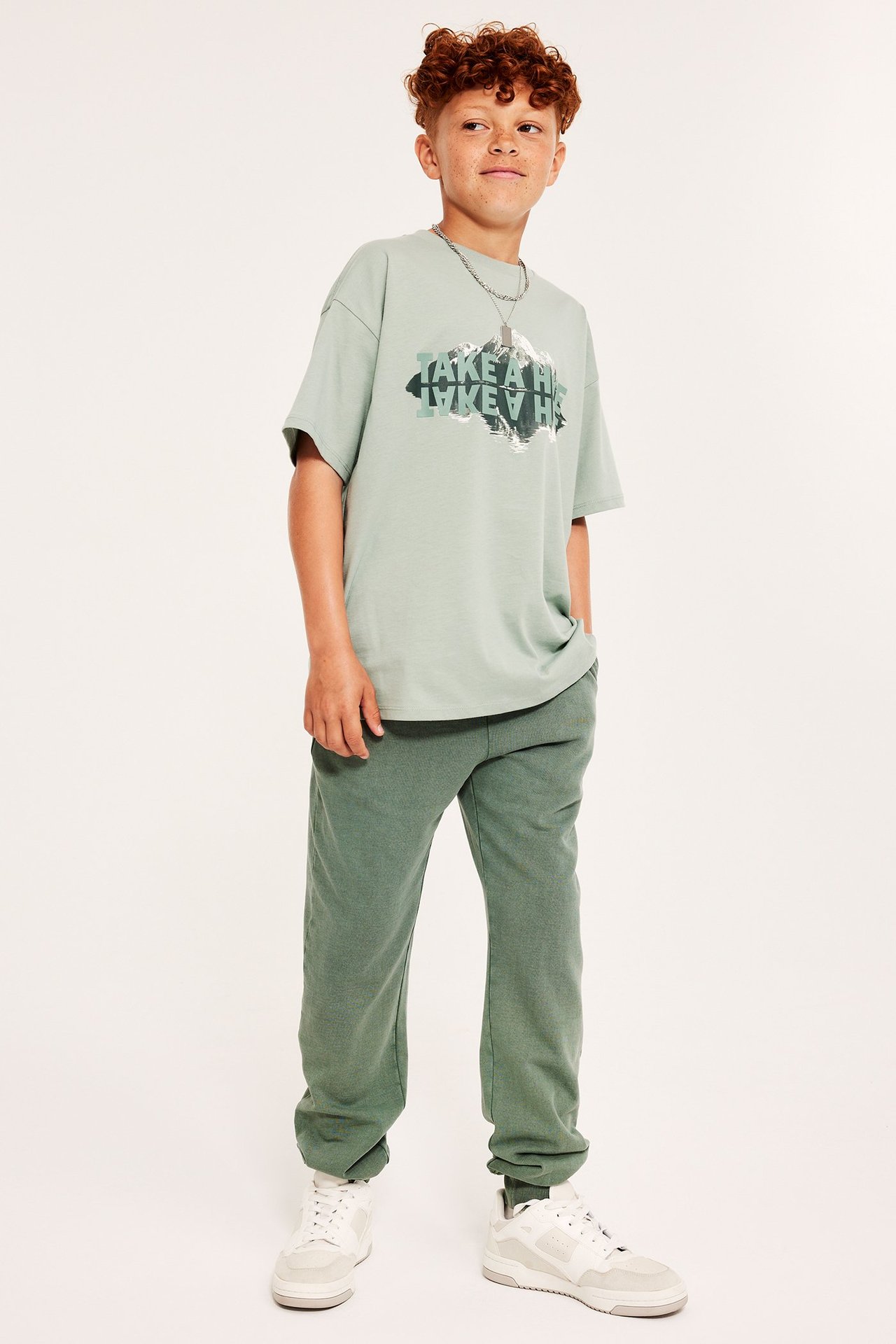 Widok z przodu: chłopiec w bawełnianych spodniach dresowych khaki, jasnozielonej koszulce z nadrukiem i białych sneakersach.