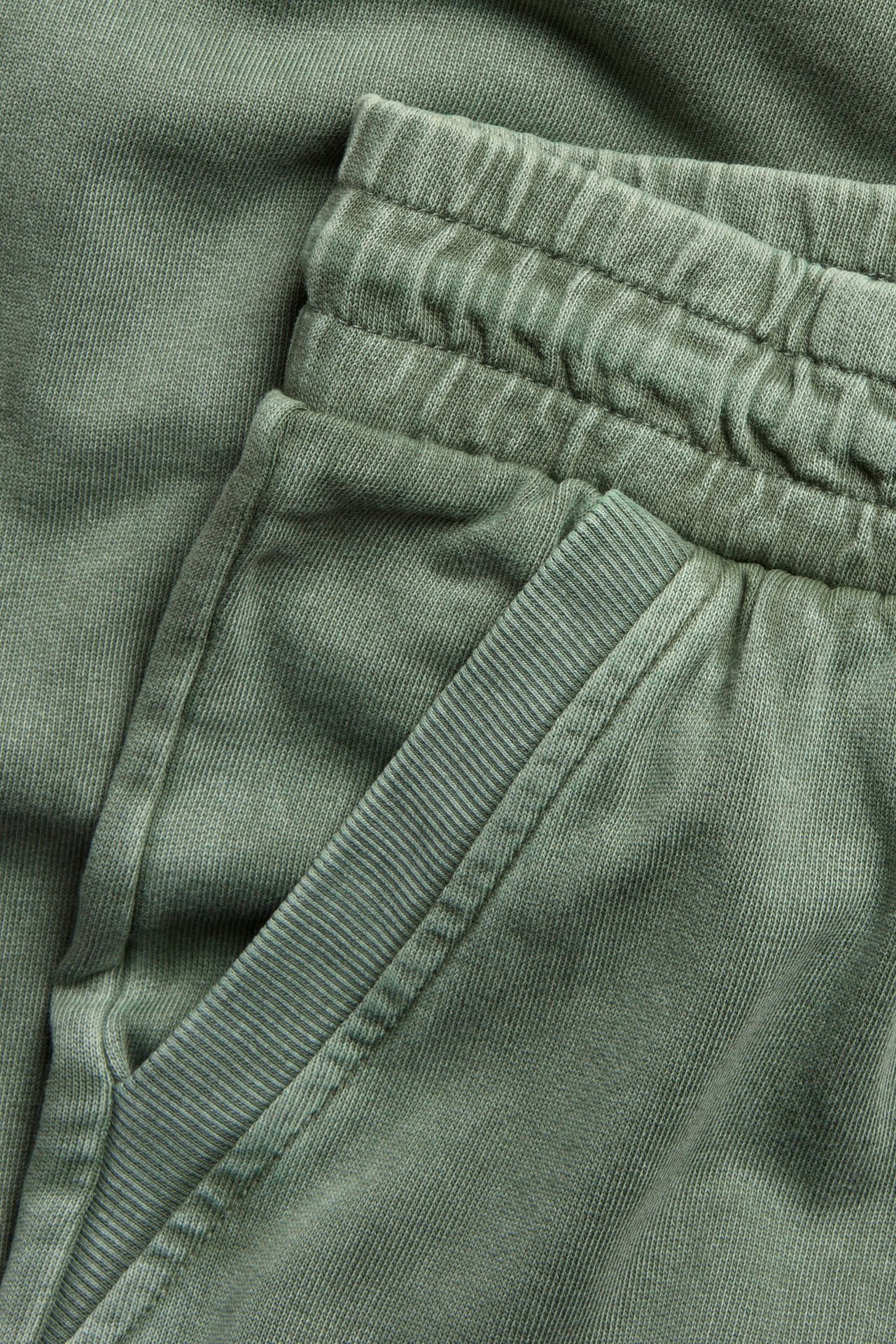 Zbliżenie na bawełniane spodnie dresowe w kolorze khaki z efektem sprania, elastyczną talią i prążkowaną kieszenią.
