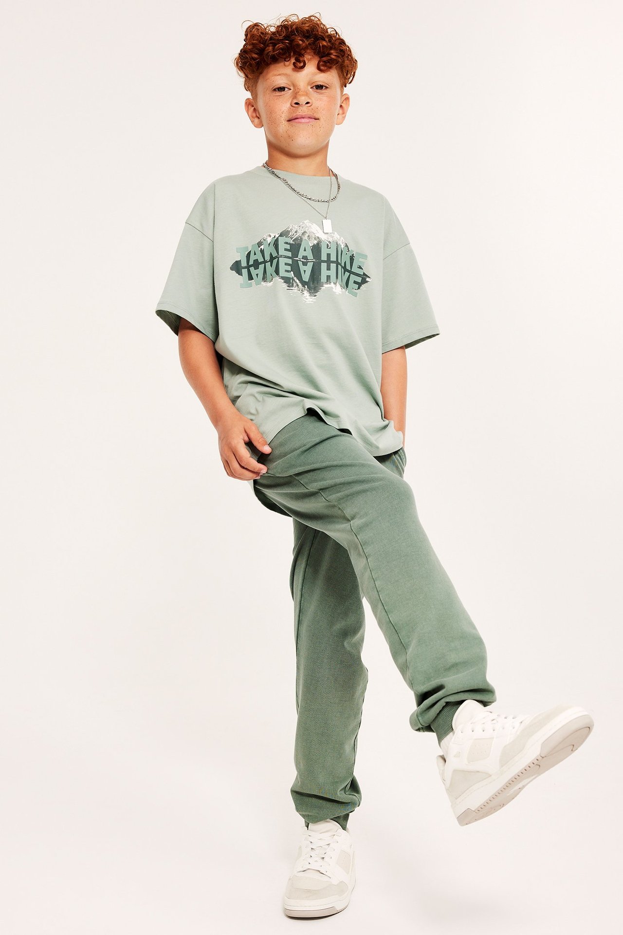 Widok z przodu: Dziecko w dresach khaki, jasnozielonej koszulce, srebrnym naszyjniku i białych sneakersach.