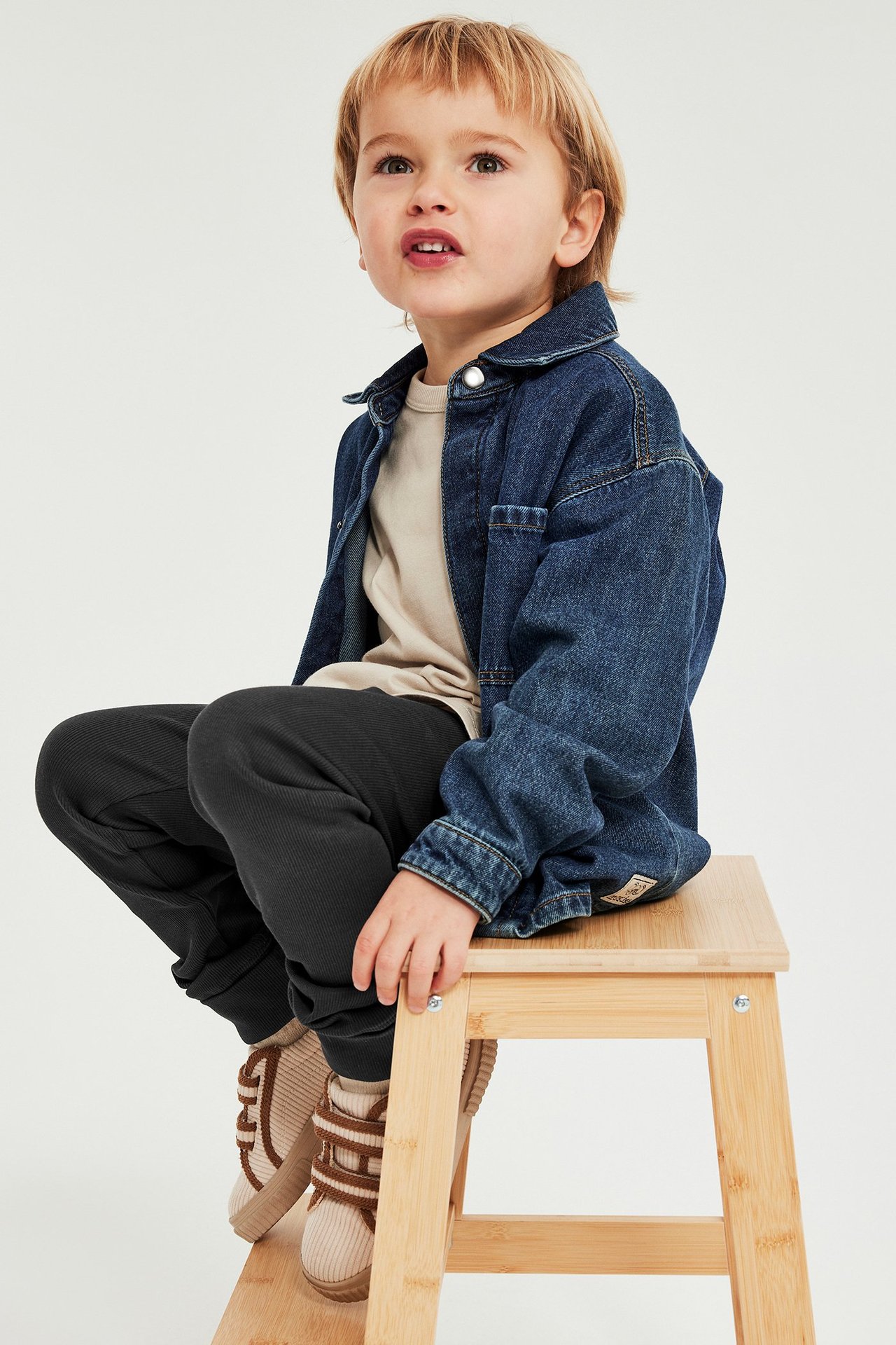 Edestäpäin: lapsi mustissa ribbileggingseissä, beigessä t-paidassa, sinisessä farkkupaidassa ja beigissä kengissä jakkaralla.