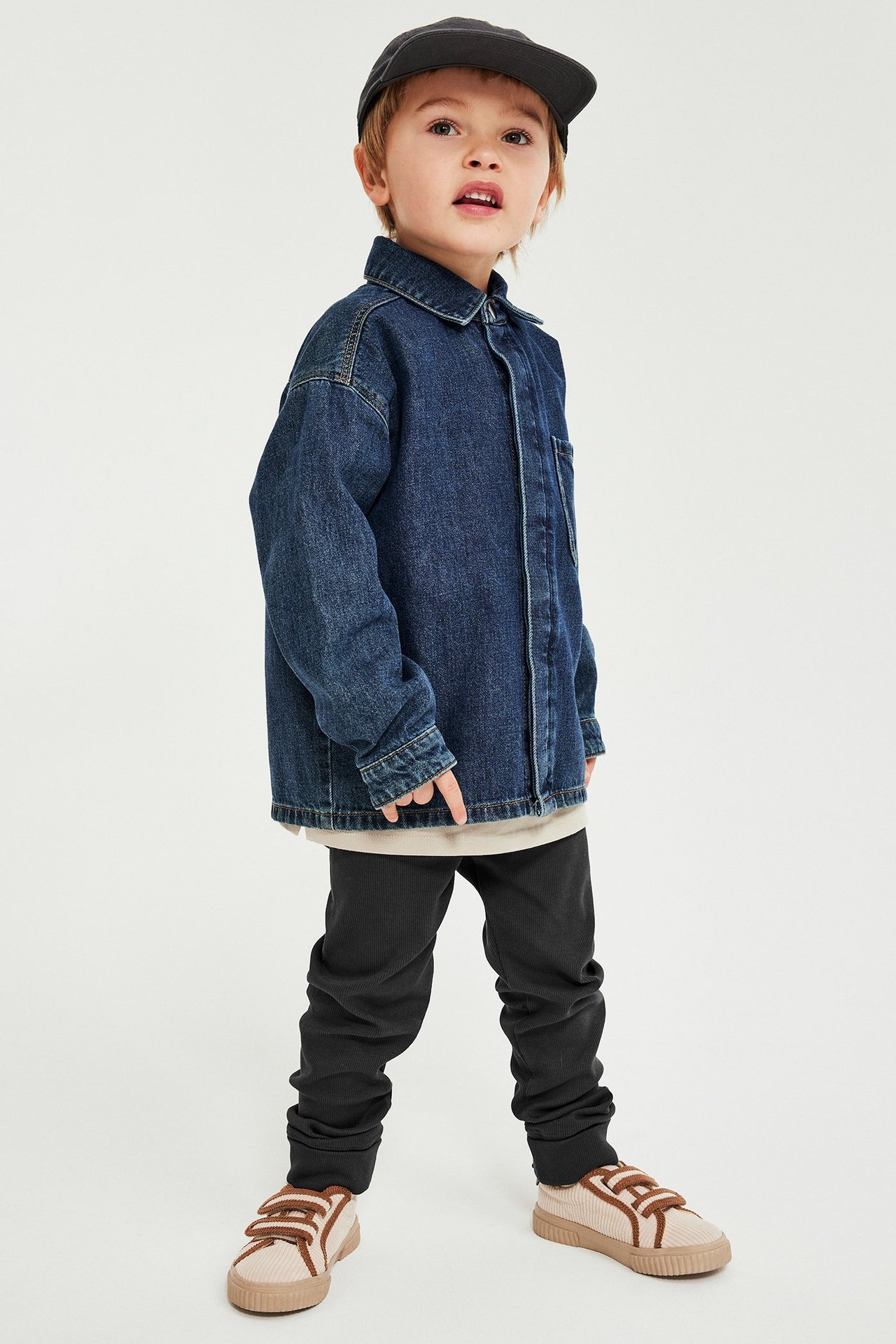 Framifrån, ett barn i svarta ribbade leggings, blå jeansskjorta, beige t-shirt, svart keps och sneakers.