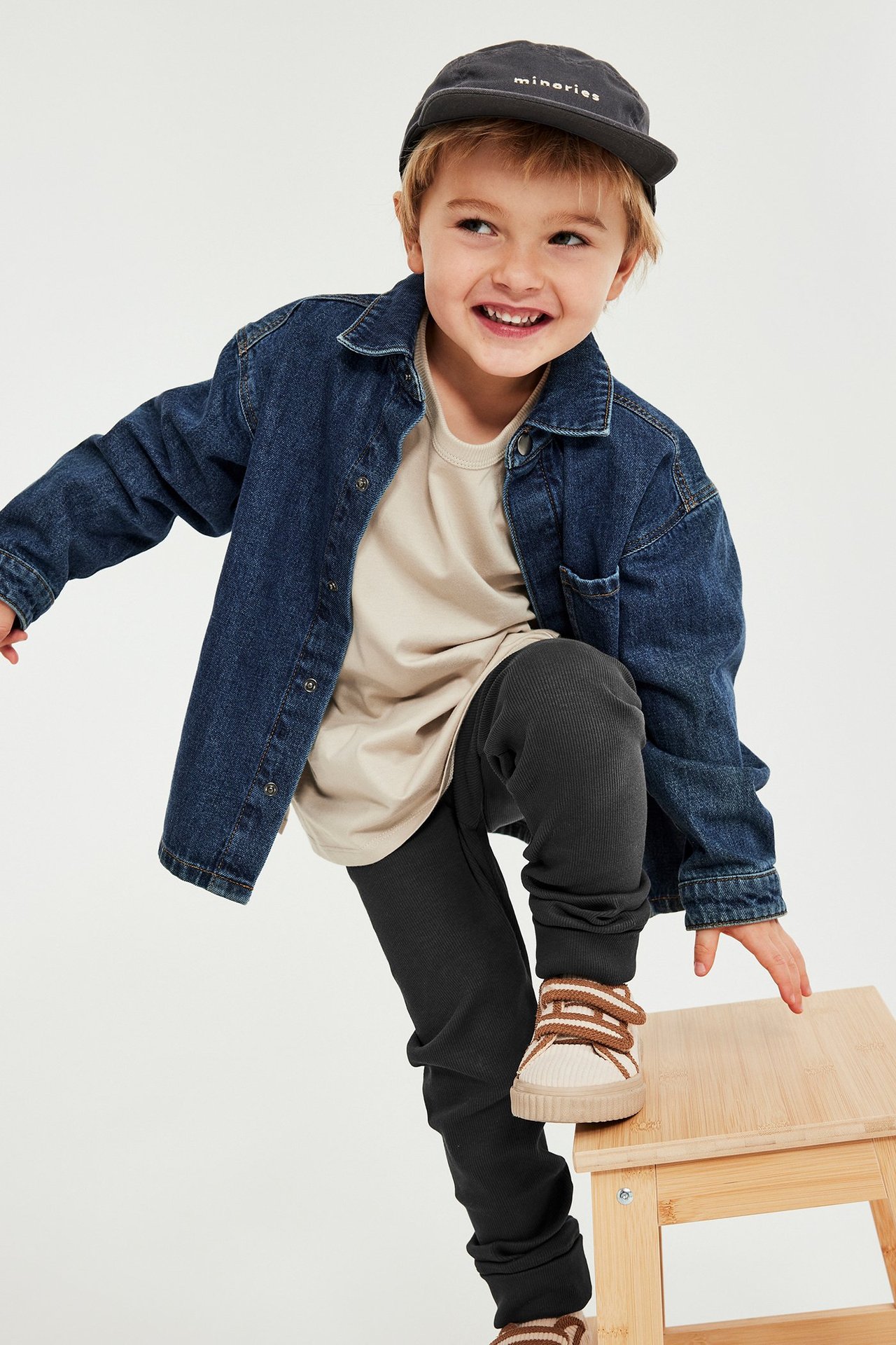 Edestä, lapsi mustissa ribbineuleleggingseissä, beigessä t-paidassa, sinisessä farkkutakissa ja tummassa lippalakissa.