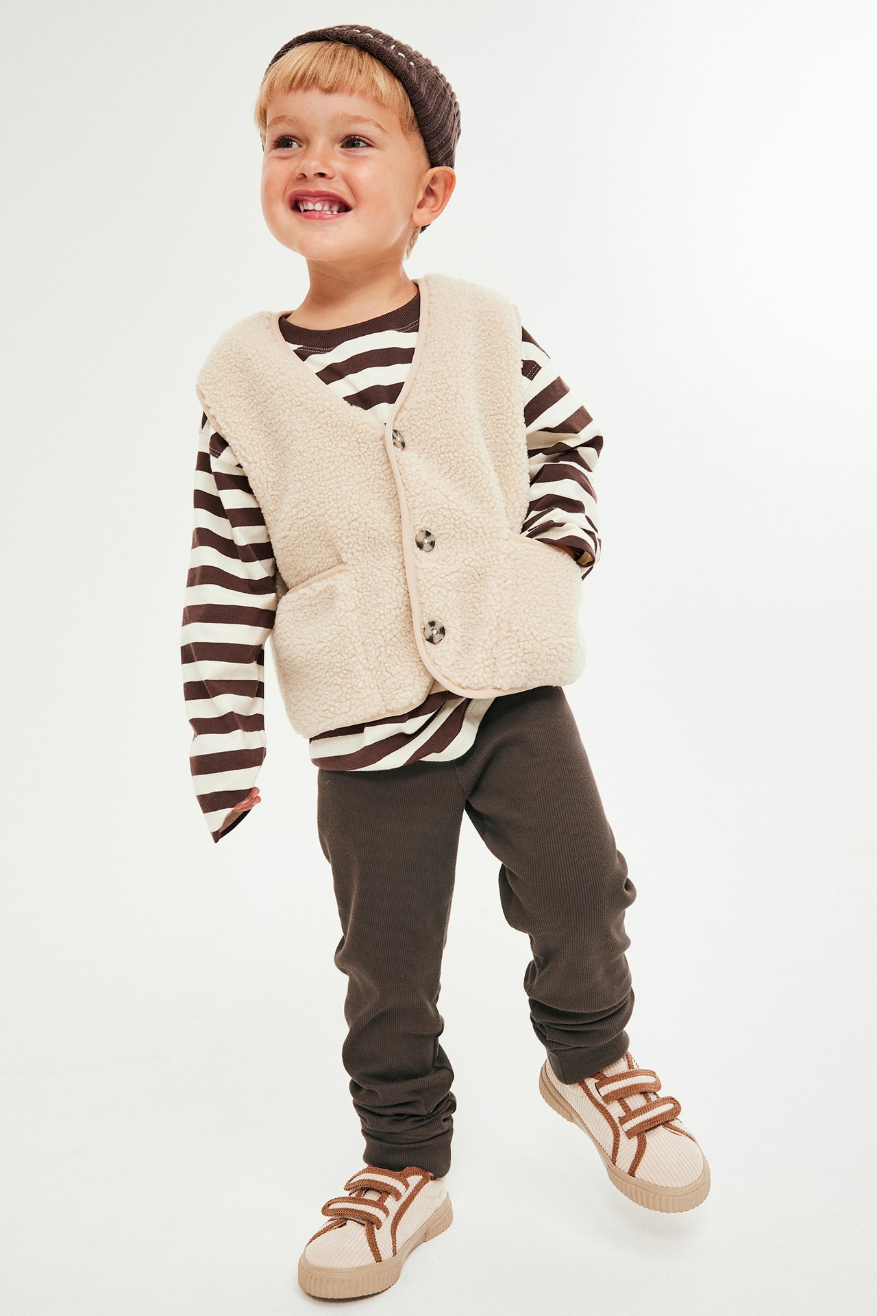 Framifrån: barn i bruna ribbade leggings, randig tröja, beige teddyväst, brun mössa, beiga sneakers.