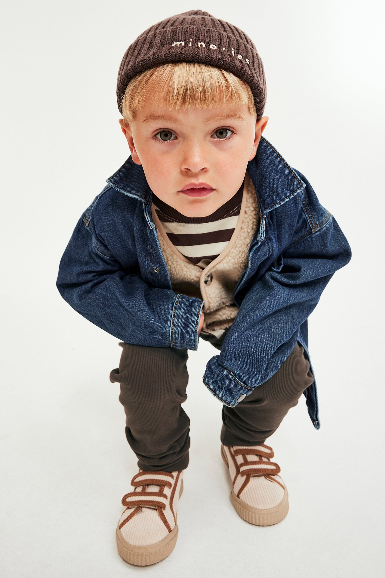 Framifrån, ett barn i bruna ribbade leggings, brun mössa, blå jeansjacka och beiga sneakers.