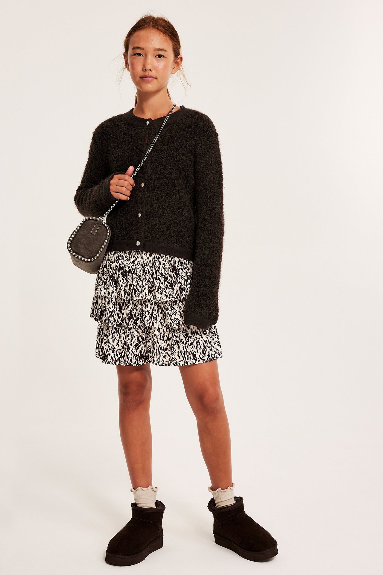 Edestä: Lapsi ruskeassa neulotussa neuletakissa napeilla, kuviollisessa hameessa, harmaassa laukussa ja ruskeissa saappaissa.