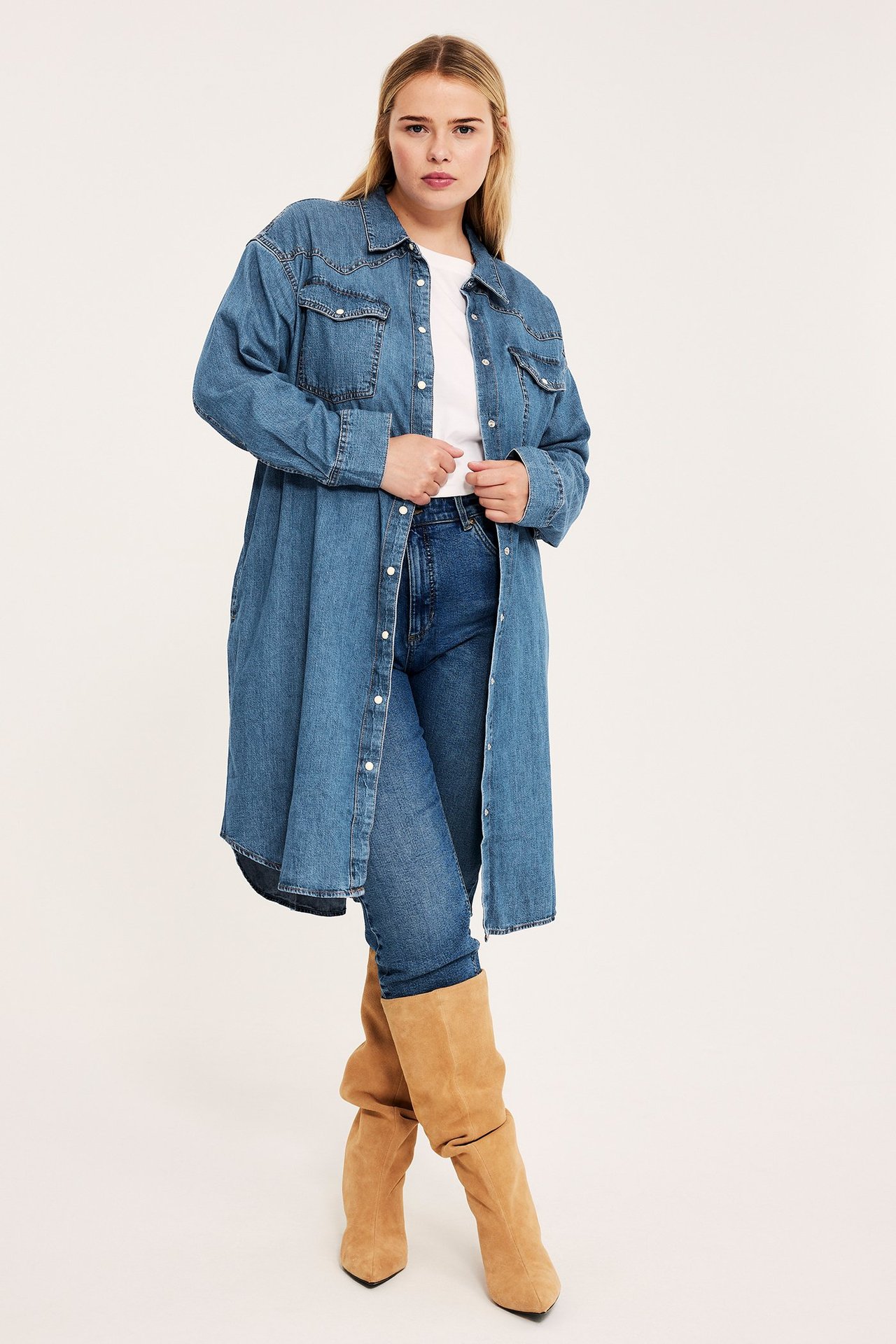 Edestä, nainen pitkässä sinisessä farkkupaitamekossa, valkoisessa t-paidassa, sinisissä farkuissa ja ruskeissa saappaissa.