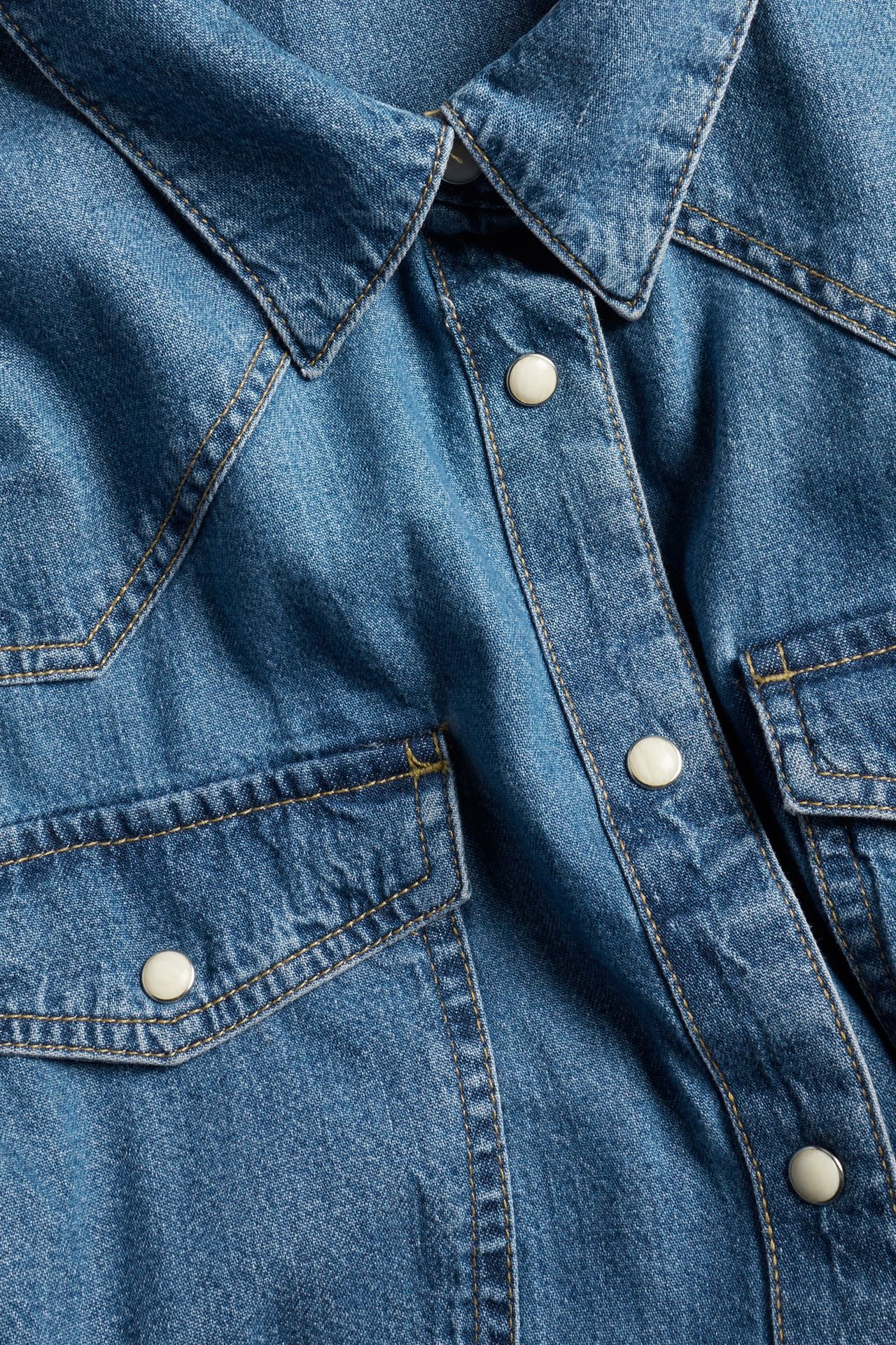 Zbliżenie na niebieską dżinsową sukienkę koszulową z kołnierzykiem, białymi zatrzaskami i dwiema kieszeniami na piersi.